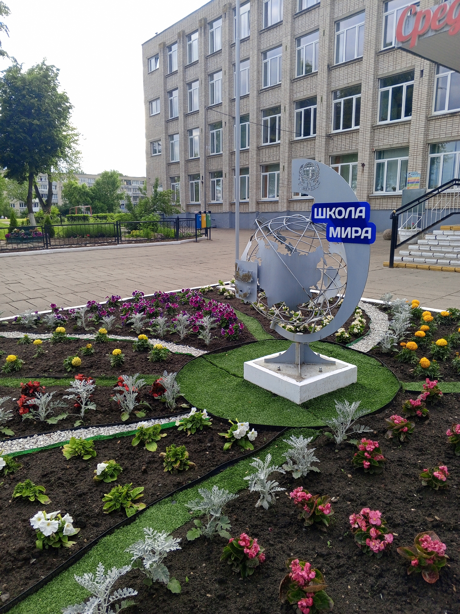 The image displays an outdoor scene dominated by a monument and a flower bed in front of a multi-story, light-colored brick building, likely a school, in the city of Unknown, Unknown. The sky is overcast.
In the foreground, a large, meticulously maintained flower bed is arranged with sections of dark soil, artificial green grass strips, and white gravel paths. Plantings include clusters of vibrant purple petunias, yellow marigolds, red and pink begonias, white petunias, and silvery-leafed plants (likely *Senecio cineraria*).
Centrally positioned within the flower bed is a monument made of grey metal. It features a stylized globe with visible latitude and longitude lines, supported by a sturdy white concrete base. Attached to the left side of the globe is a crescent-shaped panel bearing a circular emblem and the Cyrillic word "ШКОЛА" (School) below it. Two blue rectangular signs are affixed to the monument, one above the other, reading "ШКОЛА" (School) and "МИРА" (Peace) respectively, in white Cyrillic script. The monument is set on a circular patch of artificial green grass.
Behind the flower bed, a grey paved walkway extends horizontally. A black metal fence delineates the walkway from a landscaped area with trees and a distant multi-story residential building on the left. The prominent building in the background on the right is constructed of light-colored brick and features numerous rectangular windows with white frames across its multiple floors. A large, red Cyrillic sign, partially obscured, is visible on the upper right facade of this building, appearing to read "Среда" (Wednesday or Environment). A set of white concrete stairs with a black metal handrail ascends to an entrance on the building's right side. Adjacent to the stairs are a yellow and a blue trash receptacle. A smaller informational sign is mounted on the building wall near the staircase.
No individuals are present in the depicted scene.