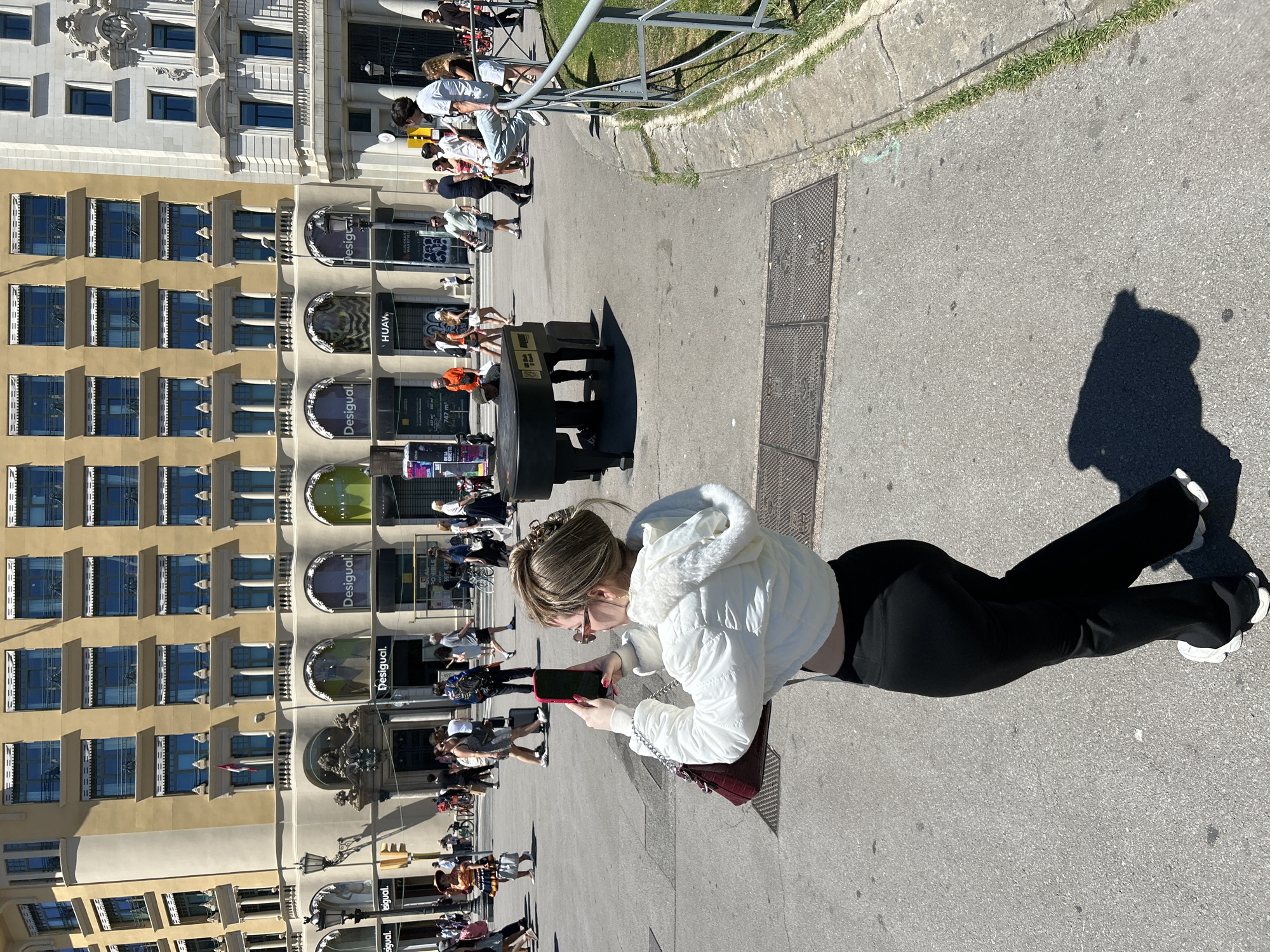 The image depicts a street scene in Barcelona, Spain, with a woman in the foreground taking a picture with her phone. She is wearing a white hooded jacket and black pants, with a red purse on the ground beside her. Behind her, pedestrians stroll past shops labeled "Desigual," "Huawei," and "Design," indicating a commercial area. A piano with bench is visible at the building. The architecture suggests a historic building with multiple floors and ornate details. The weather appears sunny, casting shadows on the pavement. A metal railing and a patch of grass are visible to the right, bordering the pedestrian area.