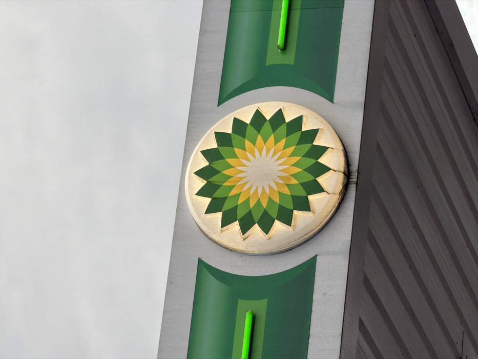 An outdoor close-up depicts the upper section of a BP gas station canopy in Vila Real, Portugal, viewed against an overcast, light grey sky. The central feature is a large, circular BP 'helios' logo, showcasing its characteristic green, yellow, and white petal design. The logo is internally lit, casting a golden glow around its perimeter, and is mounted on a light grey horizontal fascia panel. This fascia incorporates dark green accent panels with curved lighter green elements. Two prominent, horizontally aligned bright green neon or LED tube lights are integrated into these green sections, positioned to the left and right of the main BP emblem. The ribbed, grey underside of the canopy is visible in the lower portion of the frame. The entire structure exhibits visible signs of weathering and dirt accumulation. No individuals, vehicles, or active elements are present in the scene.