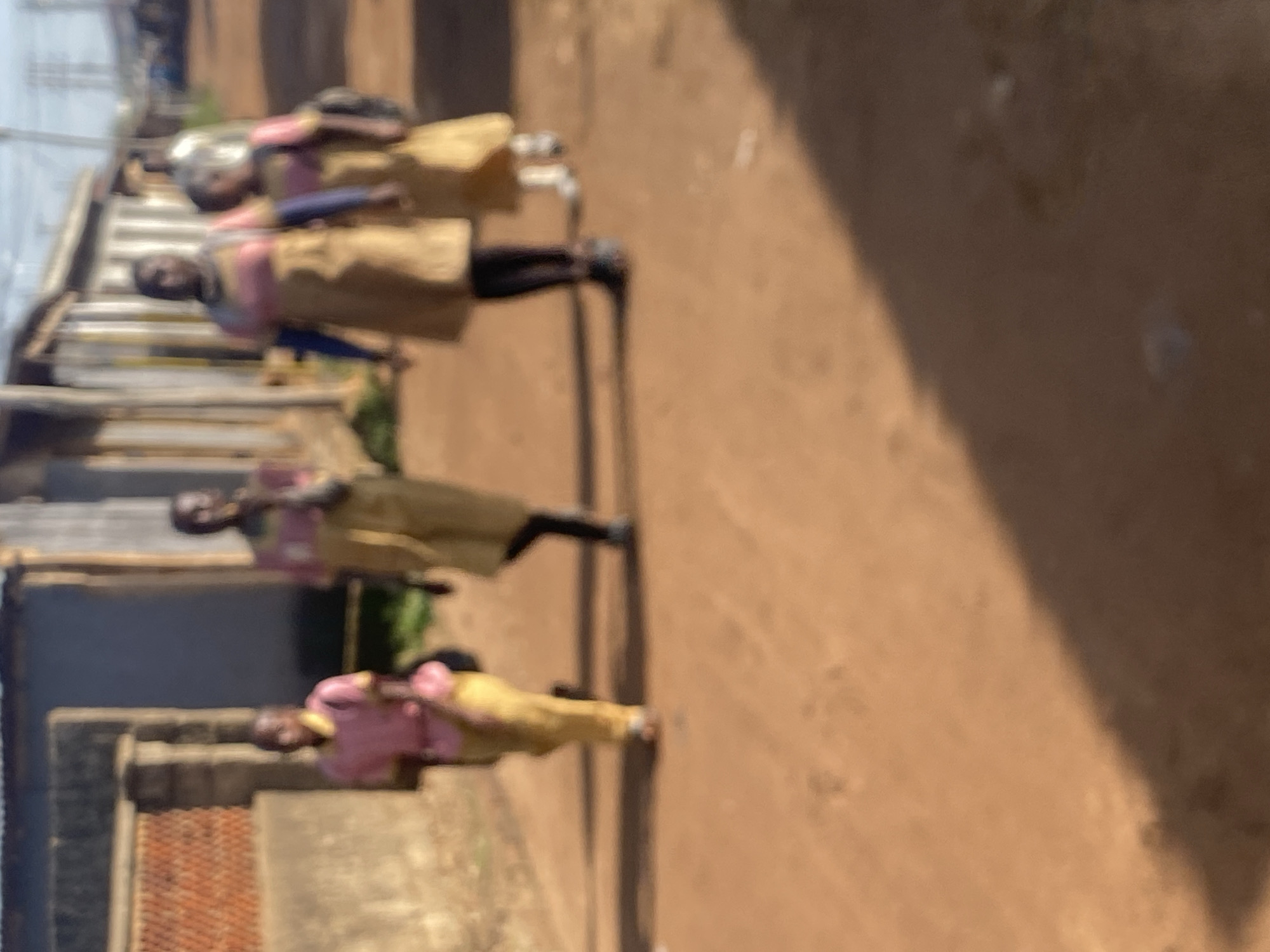 In the city of Bukuru, Nigeria, four young people, appearing to be students, are walking on a dirt road. Two individuals in the foreground are wearing light pink tops with yellow collars and yellow pants. The person on the left is carrying a pink bag. The other two individuals, further back and slightly to the right, are wearing light pink tops with yellow collars and tan or yellow dresses. The person on the far right is carrying a black backpack. The environment is characterized by a dirt road, and buildings are visible in the background, suggesting a residential or school area. The lighting indicates it is daytime.