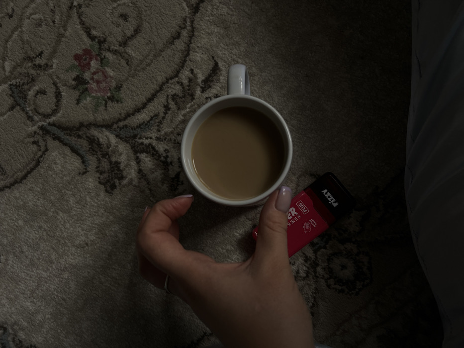 In what is identified as Leninakan, Russia, an overhead shot captures a scene of a hand, a beverage, and a vaping device resting on a patterned carpet. A light-skinned hand, with manicured fingernails painted a light lavender color and a simple silver ring on the ring finger, is positioned on the left side of the frame. The hand holds a red and black disposable vaping device labeled "FIZZY PLUS" with Cyrillic text indicating "Гранат" (Pomegranate). Immediately to the right of the hand and vaping device, a white ceramic mug filled with a light brown liquid, consistent with coffee or tea with milk, is centrally placed. The background consists of a beige or light brown carpet featuring darker grey and muted floral patterns, including small red and green accents. The lighting is dim, casting soft shadows, and a portion of what appears to be a light blue or grey garment is visible in the lower left corner.