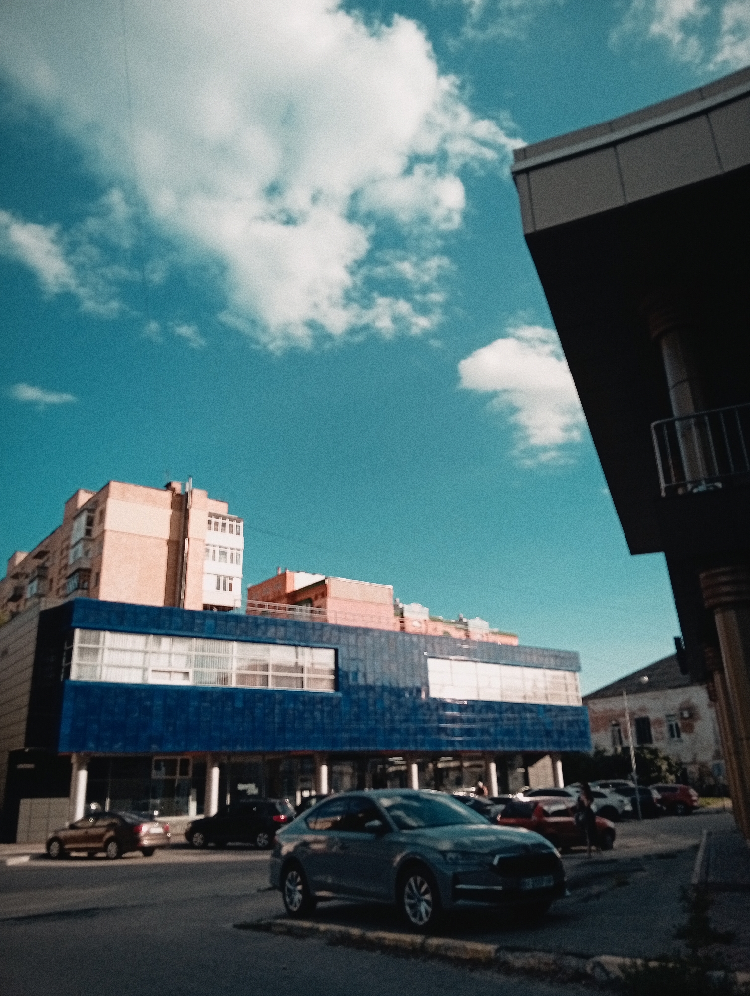 An exterior, low-angle shot captures a street scene in an urban environment under a bright blue sky with scattered white clouds. In the foreground, a light gray car is parked on the right side of the street, facing left. Behind it, other cars are parked and moving on the road. The street itself is paved, with a curb visible.
Dominating the middle ground is a modern building with a distinctive blue facade on the lower level, featuring large windows. Above the blue section, a beige, multi-story building with balconies and numerous windows rises. To the right of this building, another structure with a lighter colored facade and balconies is visible. Further in the background, a darker, more contemporary building with a large overhanging section is partially visible on the right side of the frame, extending into the foreground. Its facade appears to be dark gray with dark vertical elements. The overall scene suggests a commercial or residential area in a city.