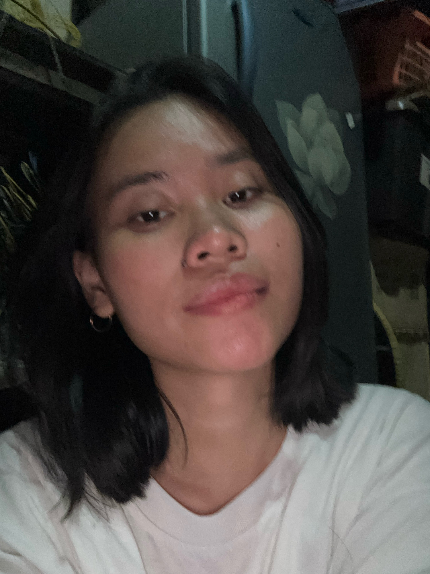 A selfie of a young woman with dark hair and light skin, wearing a white t-shirt. She has a small hoop earring in her left ear and a nose ring. Her face is slightly tilted and she is looking towards the camera. The background is dark and out of focus, with hints of objects that suggest an indoor setting, possibly a home or apartment. The lighting appears to be artificial and low-key. There is no discernible information about the location in East Jakarta, Indonesia.