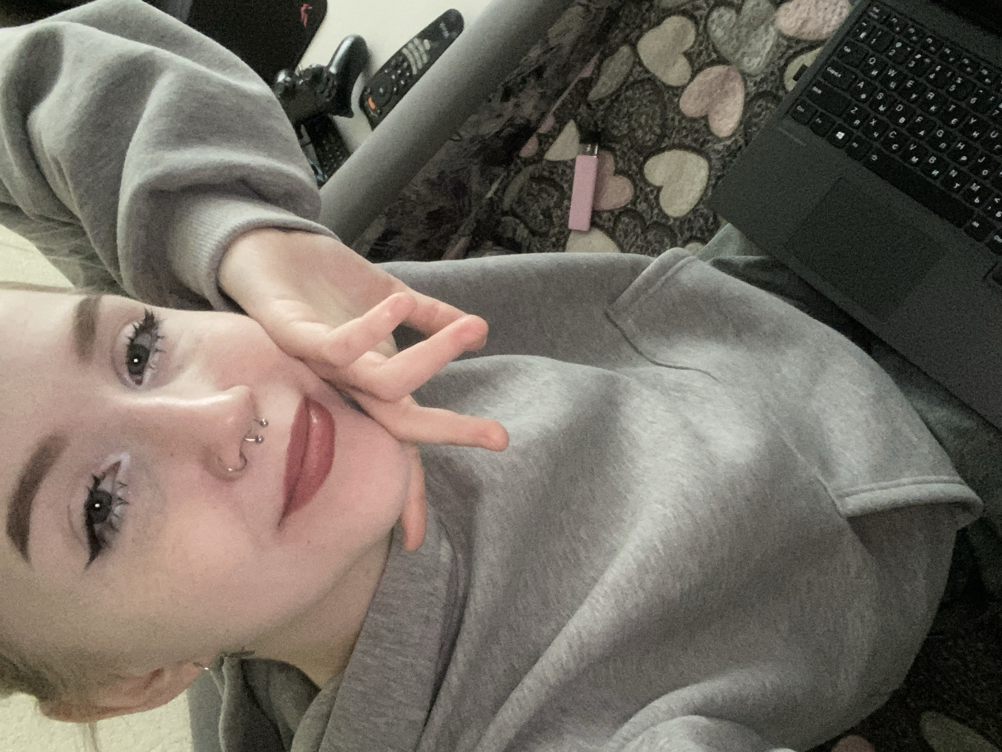 A person with fair skin, light hair, and a nose ring poses for a selfie. She is wearing a gray sweatshirt and making a peace sign with her hand near her face. The background includes a laptop, a lighter, and a patterned blanket or sheet with hearts. The environment seems to be indoors. It is not possible to confirm the location in Izhevsk, Russia, without further information.