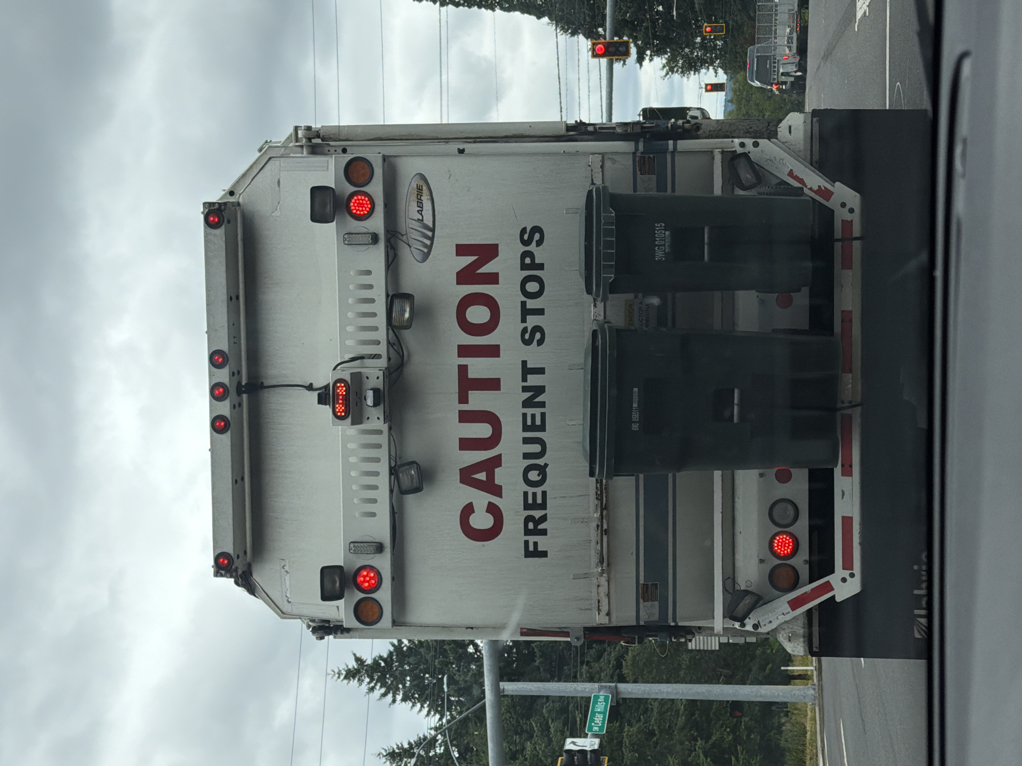 The image shows the rear of a white garbage truck with red lettering reading "CAUTION FREQUENT STOPS" on its back. The truck is equipped with two dark green garbage bins and multiple red brake lights. The truck is on a street with trees and a traffic light visible in the background. A street sign reads "SW Cedar Hills Blvd". The scene appears to be in Beaverton, United States of America. The sky is cloudy.