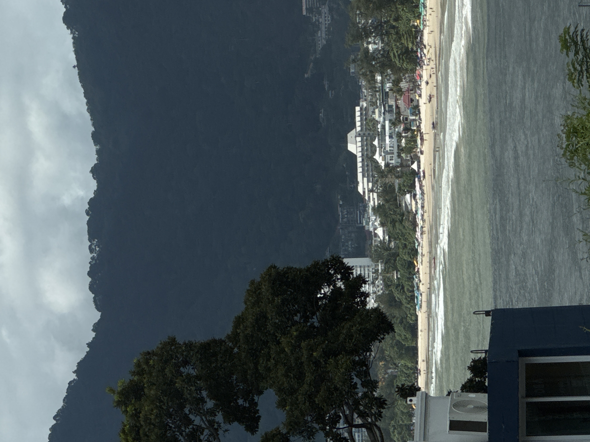 The image depicts a coastal scene in Patong, Thailand. In the foreground, a dark blue structure with a white-framed window is visible on the left, with an air conditioning unit mounted on its top. To the right, the ocean stretches out, with choppy waves rolling towards a sandy beach. The beach is lined with numerous beach umbrellas and lounge chairs, and dotted with many people. Beyond the beach, a line of white buildings, characteristic of a resort town, are clustered together. Further in the background, lush green trees and the dark, forested slopes of a mountain dominate the landscape. The sky above is overcast with dramatic, dark clouds.