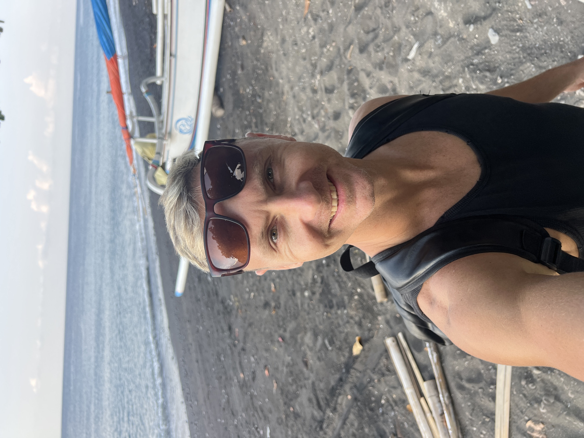 A selfie is taken by a fair-skinned man with short blond hair, wearing sunglasses with dark lenses, a black tank top, and a black backpack. He is on a beach with dark sand. In the background, the ocean is visible, with a calm sea and a clear sky. A white boat with blue and orange accents is parked on the sand. Several bamboo poles are lying on the sand to the left of the man. The scene appears to be the coast of Abang, Indonesia.