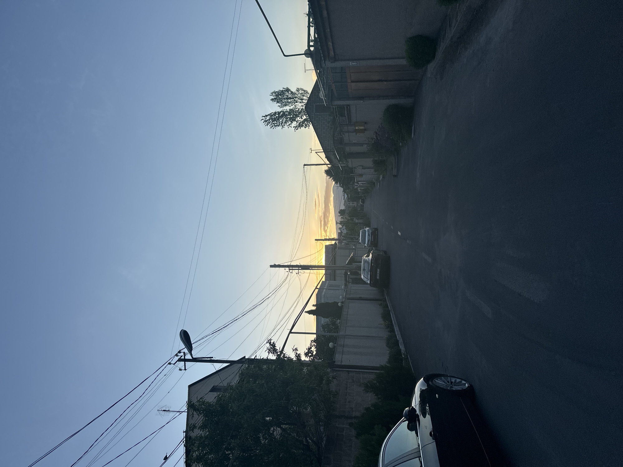 The image depicts a street scene in Arinj, Armenia. The street runs horizontally, with buildings lining both sides. Several cars are parked along the street, including a black car in the foreground. Overhead power lines crisscross the sky. The sky is light blue near the horizon and deepens to a darker blue towards the top of the frame. The sun is setting in the distance, casting a golden glow on the horizon.