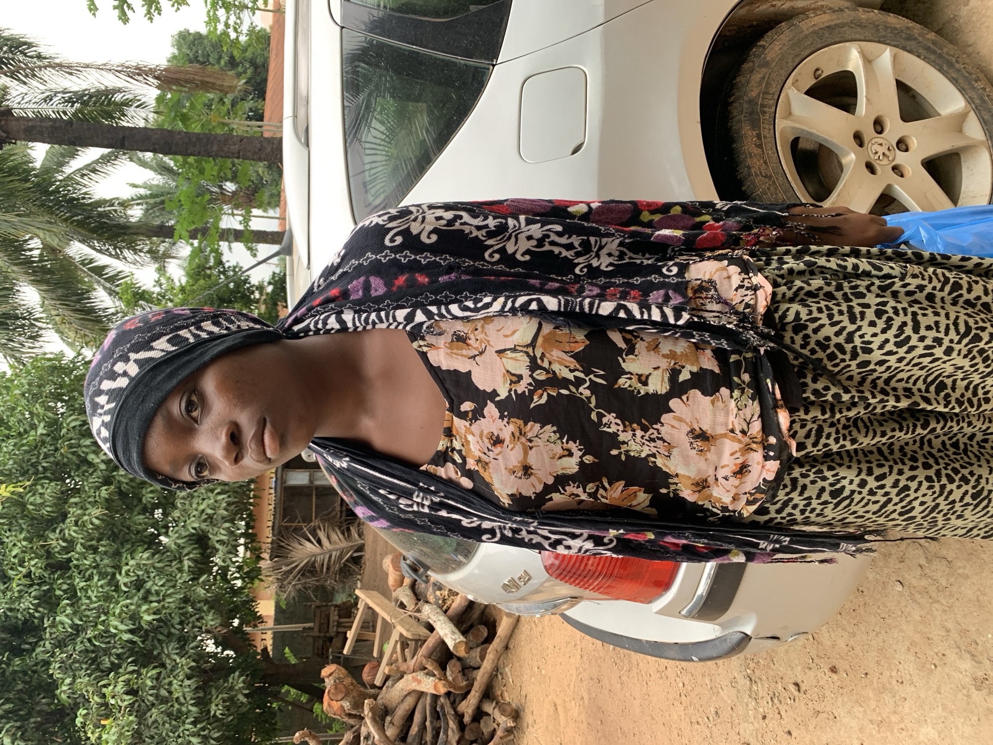 A medium shot captures a woman standing outdoors in Akwanga, Nigeria. The woman, with dark skin, looks directly at the camera with a neutral expression. She wears a black headscarf adorned with white patterns, a black floral top featuring pink and peach flowers, and a light green and black leopard print skirt or dress. Her right hand, visible on the right side of the frame, is decorated with intricate dark henna designs and holds a blue plastic bag or object.
The background features a white car, part of which is obscured by the woman. The car's rear section, including a tail light and fuel cap area, is visible, along with a dirty wheel displaying a Peugeot logo on its hubcap. Behind the car and extending into the left side of the frame, lush green trees, including tall palm trees and other dense foliage, create a tropical setting. A stack of firewood or logs is piled on the dirt ground in front of the car's rear bumper. The ground is unpaved, suggesting a rural or semi-urban environment. The lighting appears soft and diffuse, indicating either an overcast day or perhaps late afternoon. No specific activity is taking place other than the woman being present and observed.