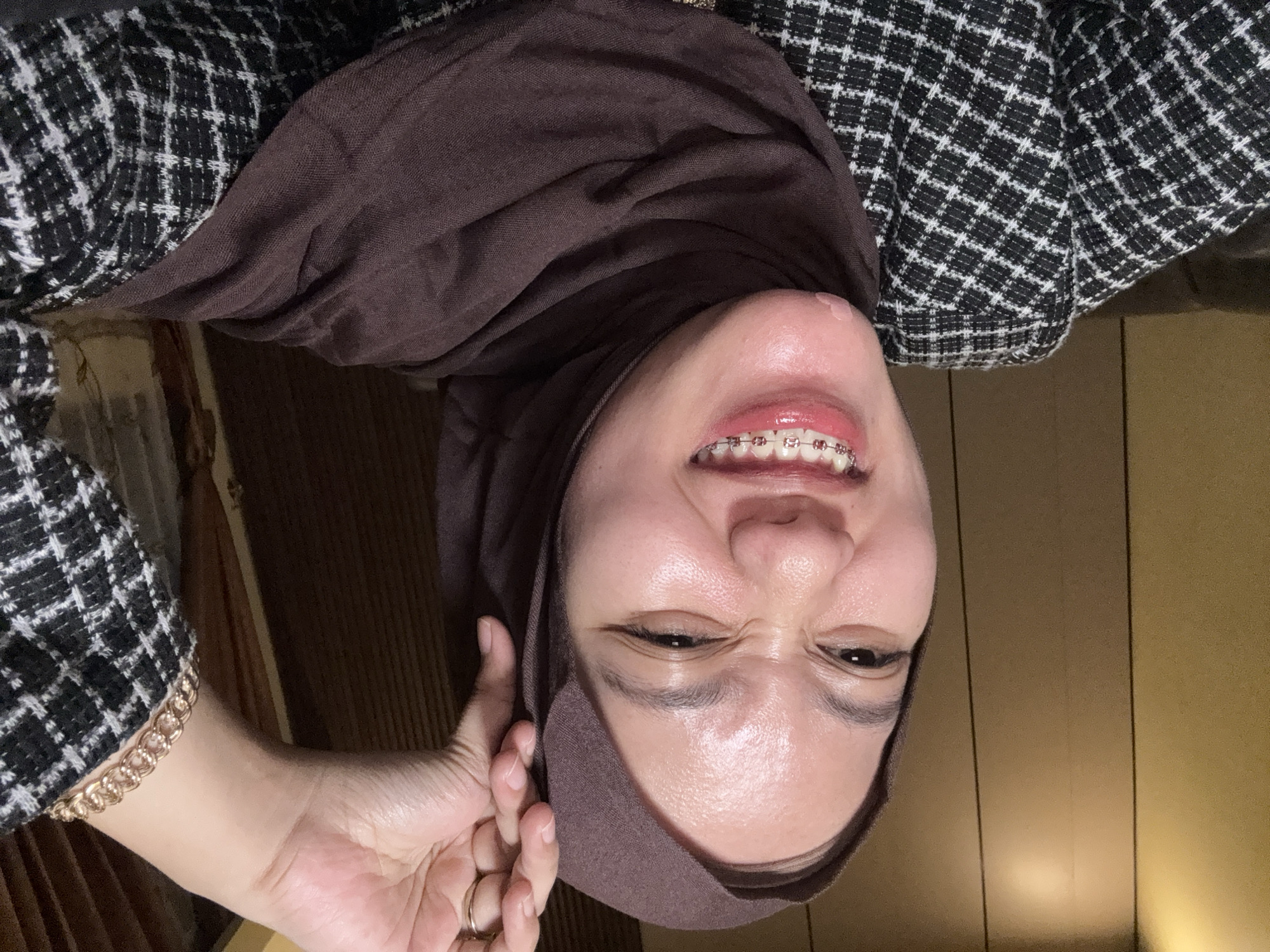A young woman wearing a brown hijab and a black and white checkered jacket smiles at the camera. She is wearing braces on her teeth. Her left hand is raised to her head. The background appears to be an indoor setting with wooden paneling. This scene is located in Gadingrejo, Indonesia.