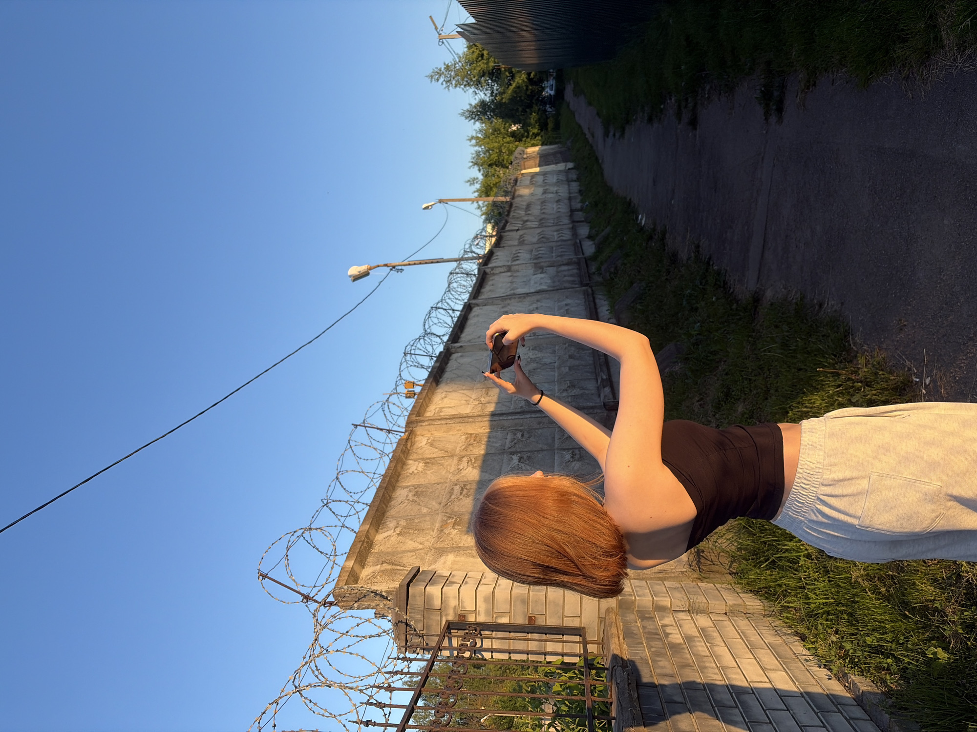 The image captures an outdoor scene in Новинки, Russia, during the golden hour, indicated by warm, low-angle sunlight and elongated shadows. A young female, seen from behind and slightly to the right, stands on the left side of a narrow, dark paved path. She has auburn bobbed hair and wears a black strapless top and light grey sweatpants with a visible pocket on the right leg. She is actively holding a dark-colored smartphone horizontally with both hands, seemingly photographing an area towards the right mid-ground.
To the subject's left, a light-colored brick pillar transitions into a dark metal fence with vertical bars, behind which green foliage is visible. Dominating the right side of the frame is a long, tall, weathered concrete wall, extending into the distance and topped with multiple coils of barbed wire. The paved path runs parallel to this wall, bordered by unkempt grass and weeds on both sides. Further along the path, two utility poles with luminaires (streetlights) are visible, and dense green trees rise above the concrete wall in the mid-ground. A single dark electrical cable crosses the clear, bright blue sky diagonally in the upper left portion of the image. Shadows from the wall and the subject extend across the ground, contrasting with the sunlit sections.