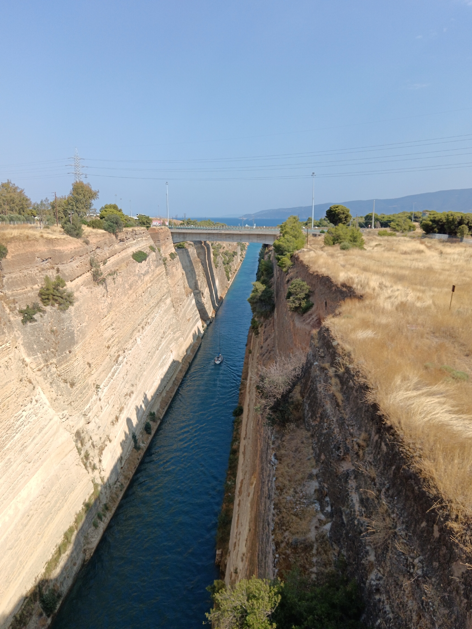 This image shows the Corinth Canal in the Municipal Unit of Loutraki - Perachora, Greece. The canal is a man-made waterway with steep, sandy-colored cliffs on either side. The water in the canal is a deep blue. A small white boat with a dark mast is navigating the canal, leaving a wake behind it. A bridge spans the canal in the background. The tops of the cliffs are covered with dry, golden grass and some scattered green trees and shrubs. The sky is clear and blue, with a few wispy clouds visible. Electrical towers and power lines are present on the land in the background, suggesting nearby infrastructure. The overall scene depicts a dramatic and engineered landscape under a bright, sunny sky.
