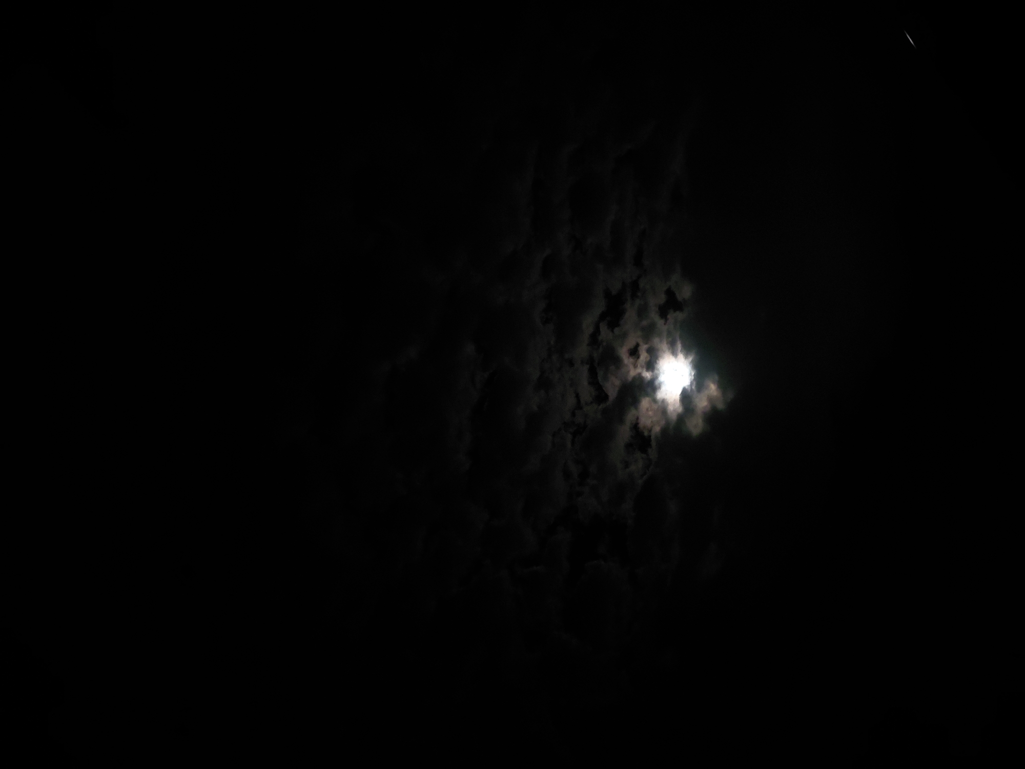 The image depicts a nighttime sky over Santa Ana, Venezuela, dominated by the moon partially veiled by clouds. The moon, positioned slightly to the right of the center, emits a bright, intense light that illuminates the surrounding dark, dense cloud formations. The clouds appear textured and broken, allowing the moon's glow to pierce through gaps and outline their edges.
The majority of the sky is deep black, emphasizing the stark contrast with the moon's radiance and the lighter shades of the illuminated clouds. In the upper right corner, a faint, thin, white linear streak is visible against the dark sky, possibly indicating the trajectory of an aircraft or a satellite. The scene is devoid of any specific terrestrial features or human-made objects other than the potential aerial streak. No text is visible in the image. The setting is clearly a cloudy night, with the moon acting as the primary light source.