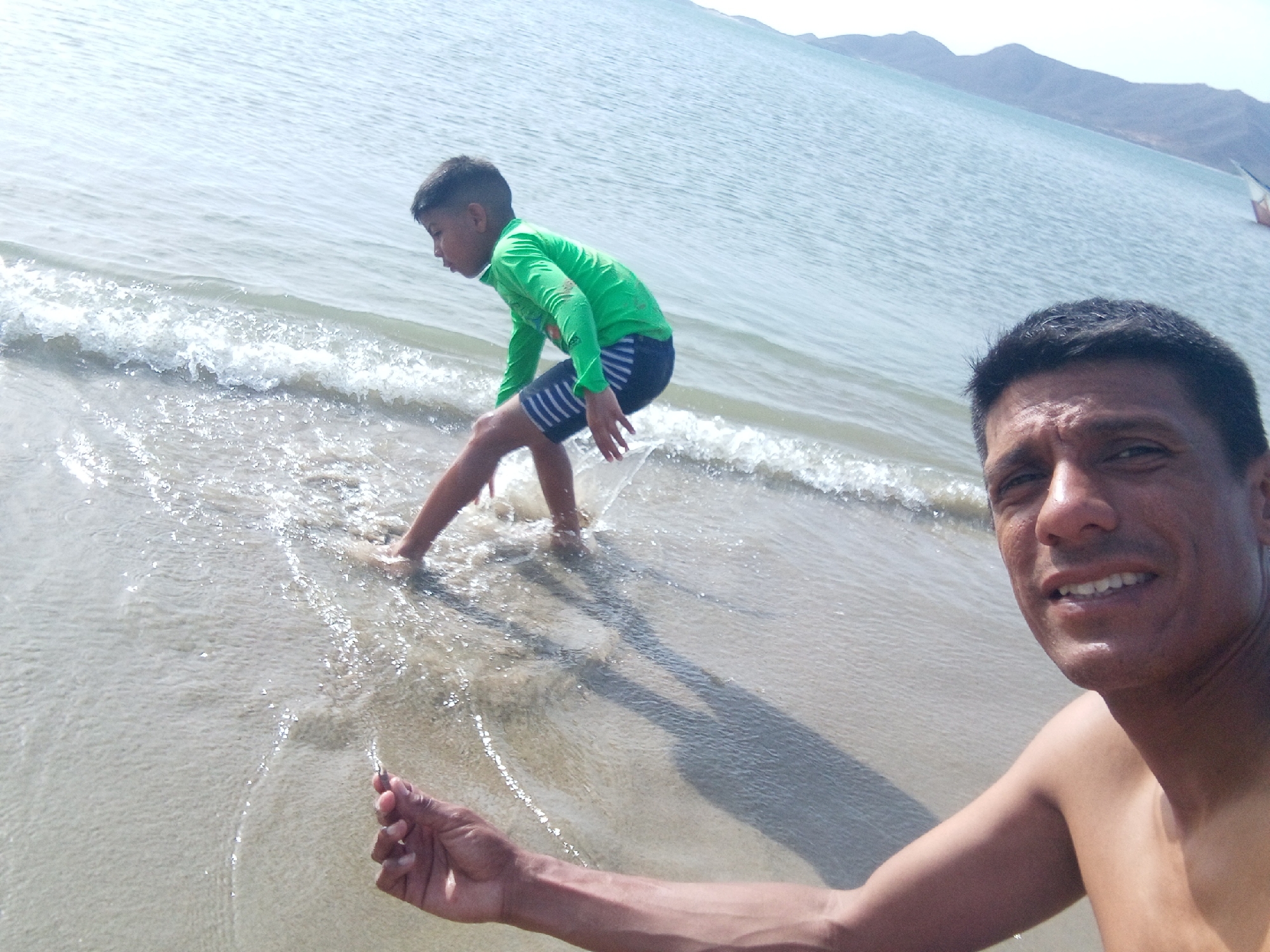 A medium shot taken at what appears to be Playa La Galera, Juan Griego, Venezuela, shows an adult man with a bare chest and dark hair at the right of the frame, smiling. In the background to the left, a young boy wearing a green long-sleeved rash guard and blue striped swim trunks is standing in the shallow water, seemingly testing the water. The beach's sand is a light tan, and the water is clear with small waves. In the far distance, there are mountains, and a boat is barely visible. The lighting is bright, suggesting it's daytime.