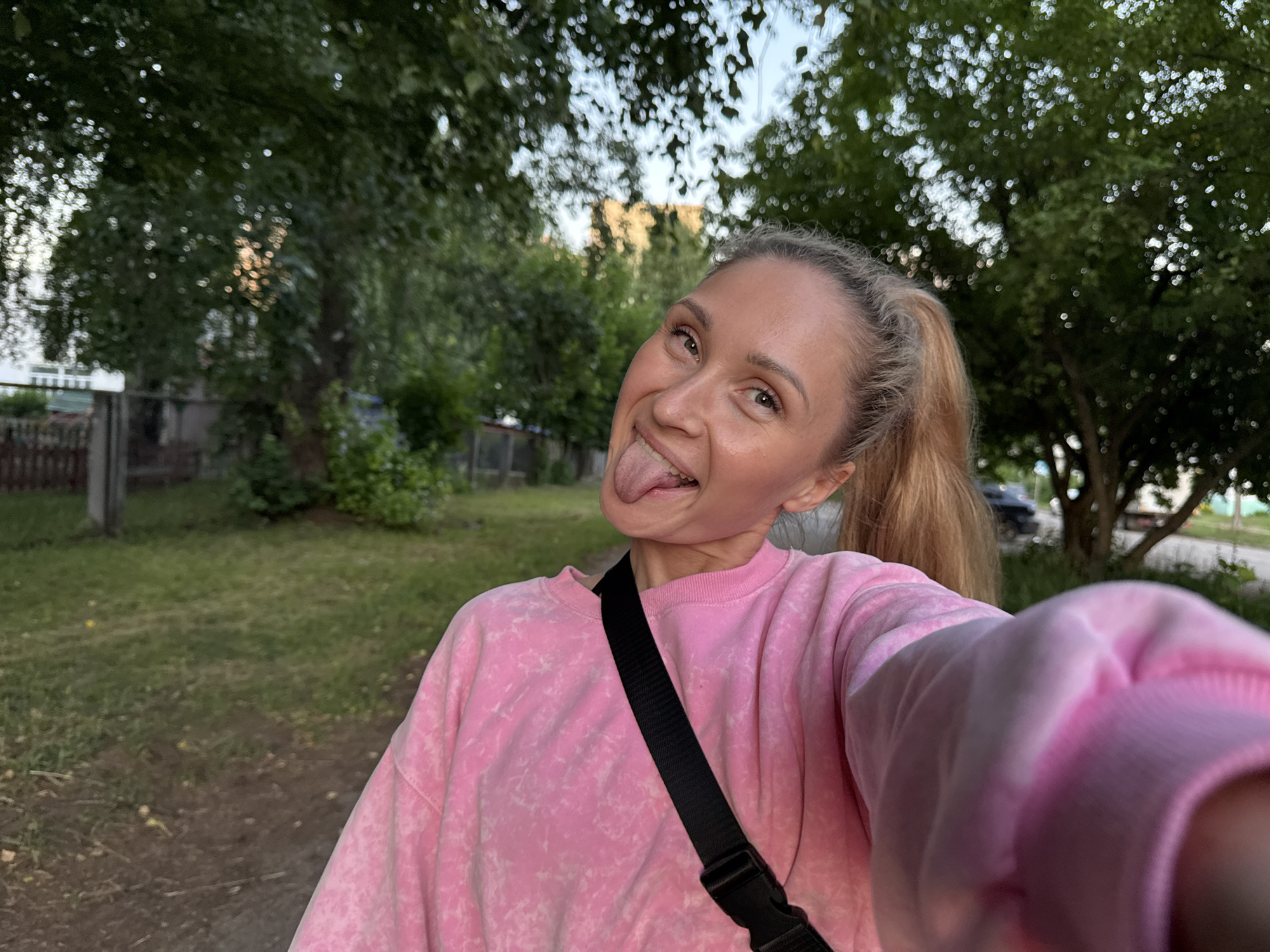 A young woman with blonde hair pulled into a ponytail, wearing a pink tie-dye sweatshirt and a black strap across her chest, is sticking her tongue out at the camera. She is outdoors, in what appears to be a park or residential area with trees, grass, and a fence visible in the background. The setting is consistent with a location in the city of Chaikovsky, Russia, given the typical residential and park-like environments found there. The lighting suggests it is either early morning or late evening.