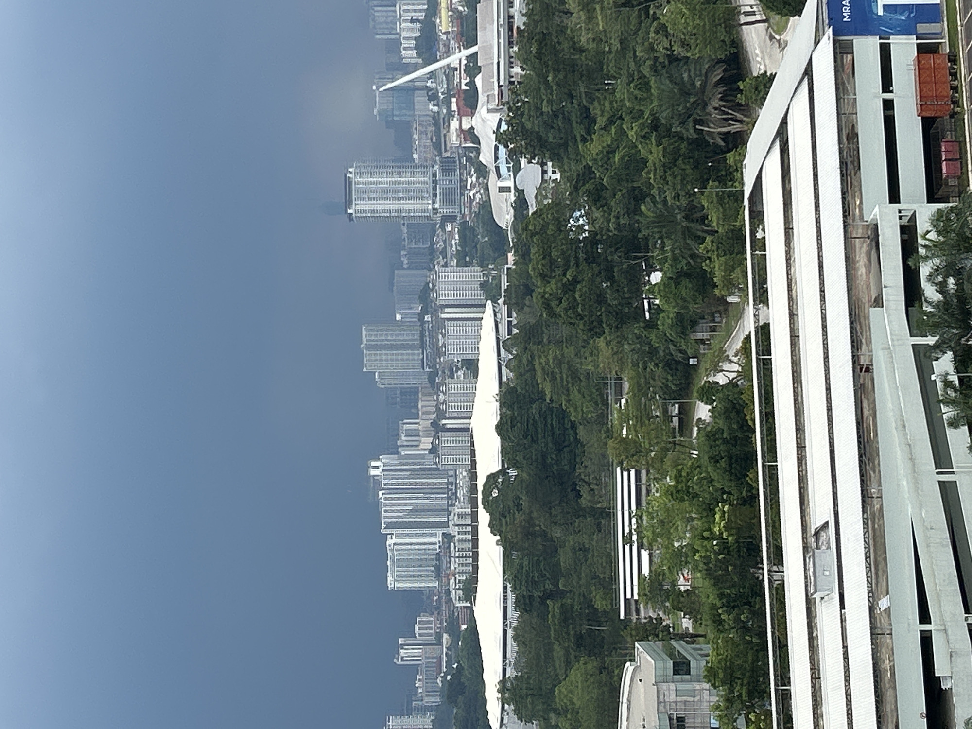 The image shows a view of the city of Kuala Lumpur, Malaysia. Tall residential buildings are densely packed along the coastline. A large building with white vertical stripes is in the foreground, partially obscuring a view of greenery and streets. The sky is overcast, with a dark bluish-gray hue filling the left portion of the image.