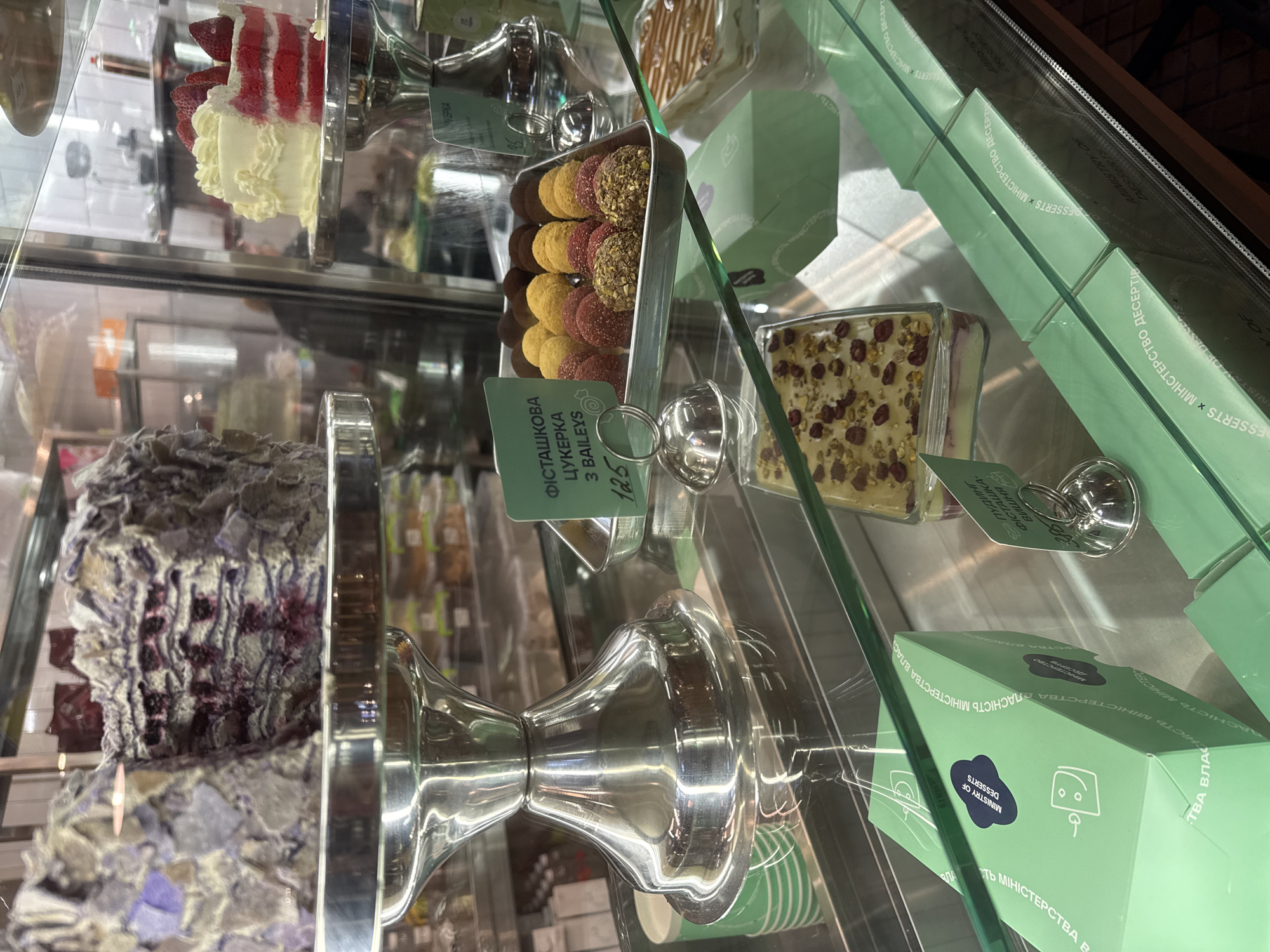 Here is a forensic description of the image:
**Scene:** Interior shot of a pastry display case in a retail establishment.
**Objects:**
* Variety of cakes and confections, including:
* A slice of red velvet cake with white frosting and strawberry slices.
* Cake of shredded chocolate.
* Tray of spherical candies in different colors.
* Dessert dish containing custard-like filling.
* Silver-colored stands and serving dishes for the pastries.
* Green boxes labeled "MINISTRY OF DESSERTS" in English.
* Price tags.
**Environment:**
* Glass display case.
* The desserts are for sale.
* The image captures a commercial setting focused on pastries.
**Text Elements:**
* Various lines of text on the boxes and price tags in Ukrainian.
* The labels contain words such as "МІНІСТЕРСТВО" (Ministry).
* Price "125" is visible on a price tag.