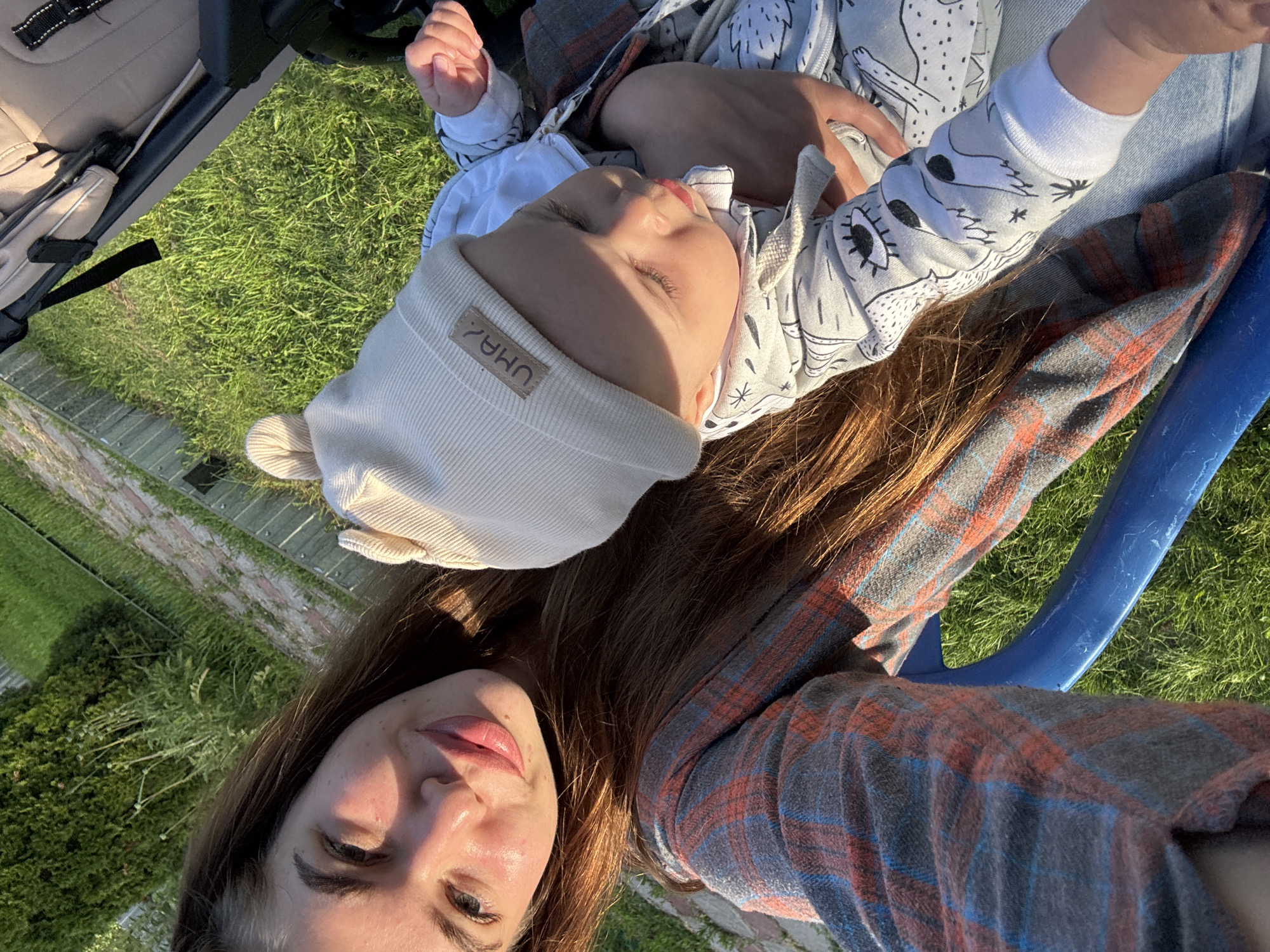 The image shows a woman and a baby outdoors, with grass and what appears to be a park or backyard in the background. The scene is bathed in the warm light of what seems to be late afternoon or early evening. The woman is in the foreground, looking towards the camera with a gentle smile. She has long, brown hair and is wearing a plaid shirt. The baby is lying on the woman's chest or lap, facing away from the camera and looking towards the left. The baby is wearing a white beanie with a small label that appears to have some text on it, possibly "UM&?" and a cartoonish design on its onesie. The overall mood of the image is peaceful and affectionate, suggesting a moment of quiet bonding between mother and child. The lush green grass and the golden sunlight indicate a pleasant, likely warm, day. The text "UM&?" is visible on the baby's beanie.