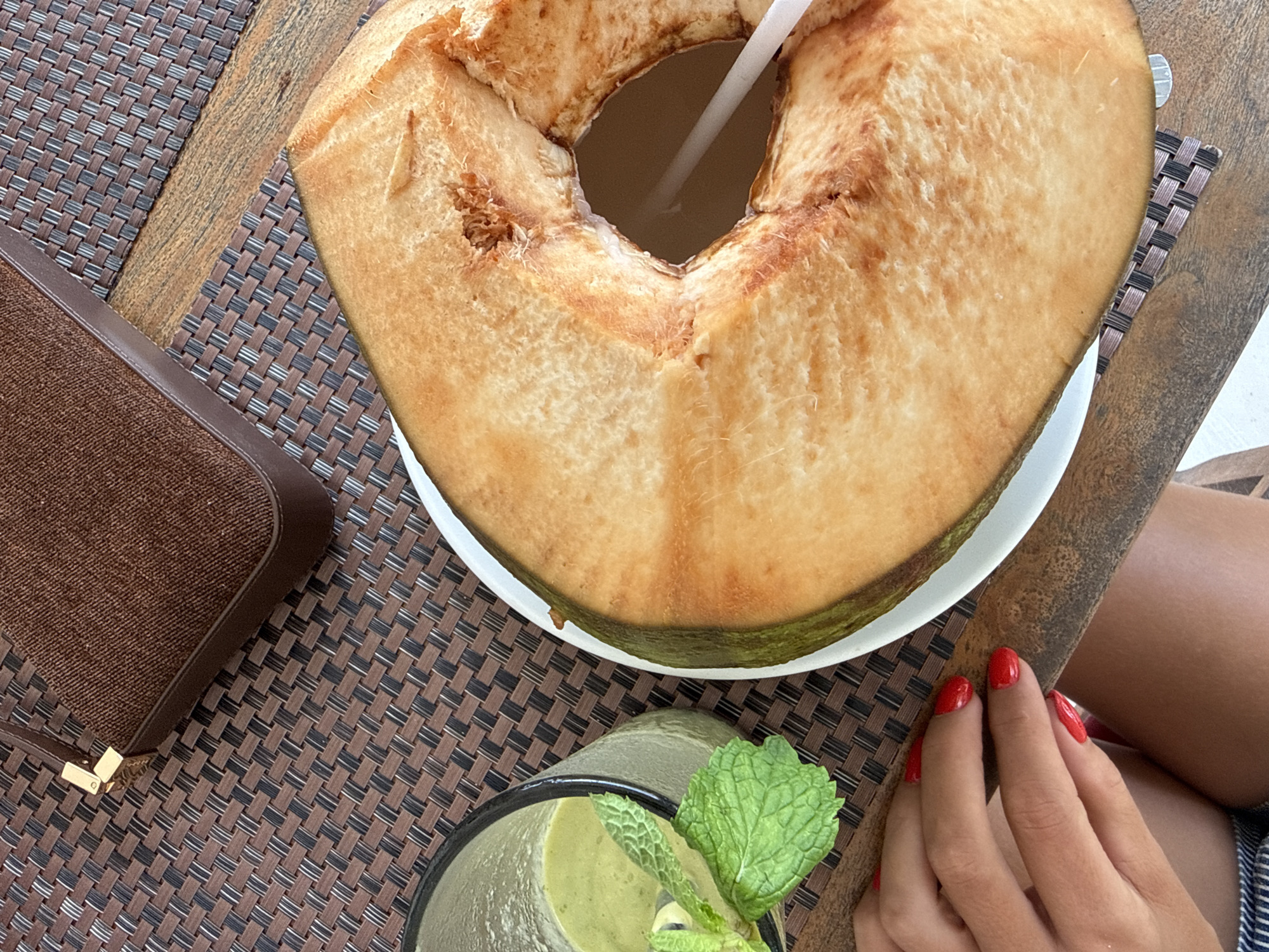 A high-angle shot shows a table in Kuta Selatan, Indonesia, arranged with food and personal items. Dominating the scene is a halved coconut on a white plate, containing a straw for drinking. Adjacent to it is a glass of green smoothie, garnished with mint leaves. A brown purse sits on the table's edge, while a woman's hand with red nail polish rests on the table. She is wearing blue and white striped shorts. The table features a woven brown placemat over a wooden surface.