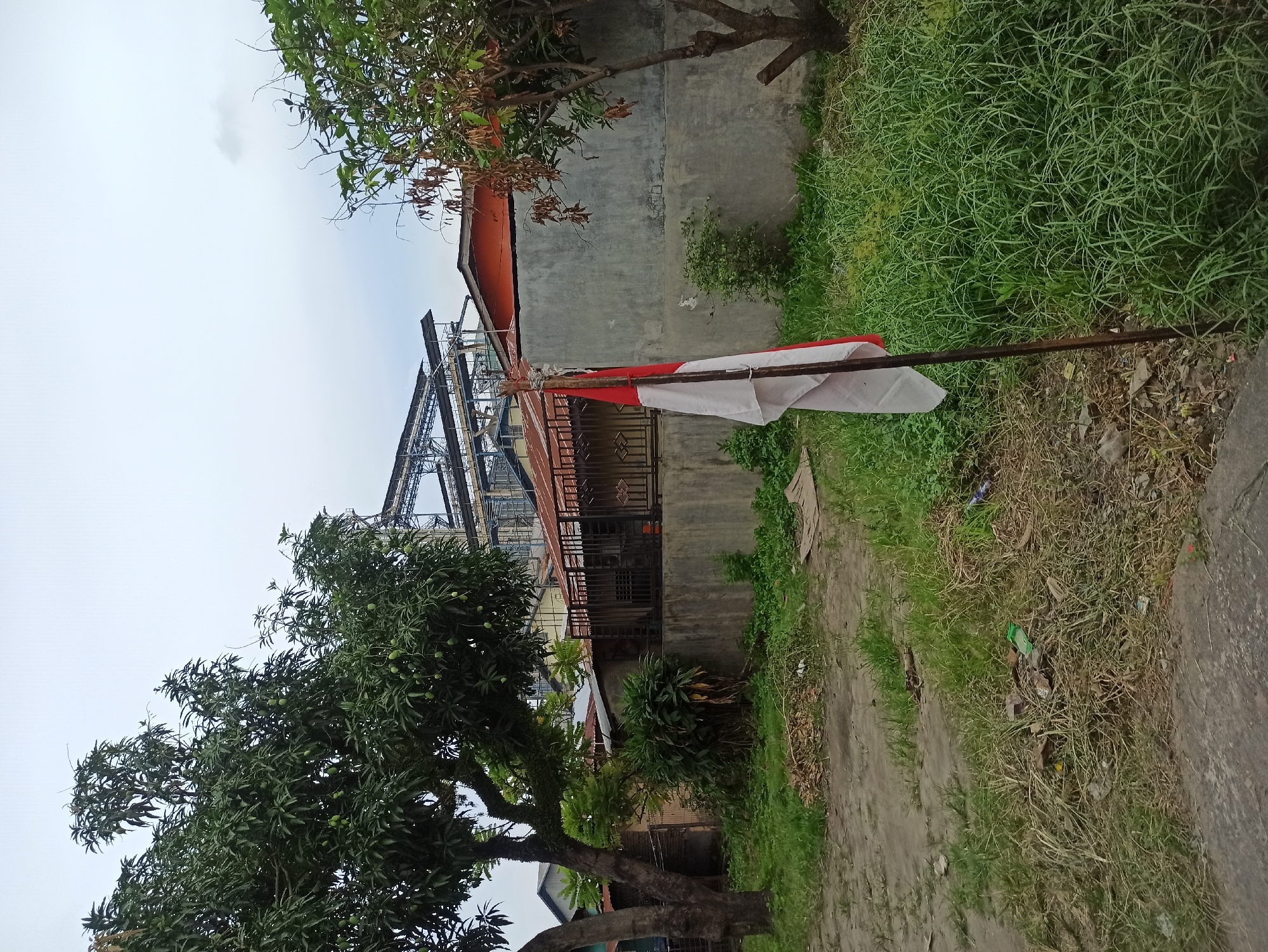 The image depicts a slightly overcast outdoor scene with an Indonesian flag planted in the foreground on a metal pole. The flag, which is red and white, is partially unfurled and appears somewhat weathered. To the left of the flag, a dirt path is visible, bordered by dry leaves and sparse vegetation.
Behind the flag and the path, there is a concrete wall with an orange-tiled roof visible above it. Beyond the wall, a complex industrial structure with metal scaffolding and piping is prominent in the background. This structure suggests the presence of a factory or processing plant.
To the left of the industrial structure, trees with dense foliage, possibly mango trees with small green fruits, are present. The general environment appears to be an urban fringe area, where residential or older structures meet industrial zones. The ground is covered with a mix of grass, dirt, and some scattered debris, including a plastic bottle. The sky is a uniform pale gray, indicating cloudy weather. The specific location within the City of Medan, Indonesia is not explicitly identifiable from the visual cues alone, but the presence of the Indonesian flag strongly suggests the country.