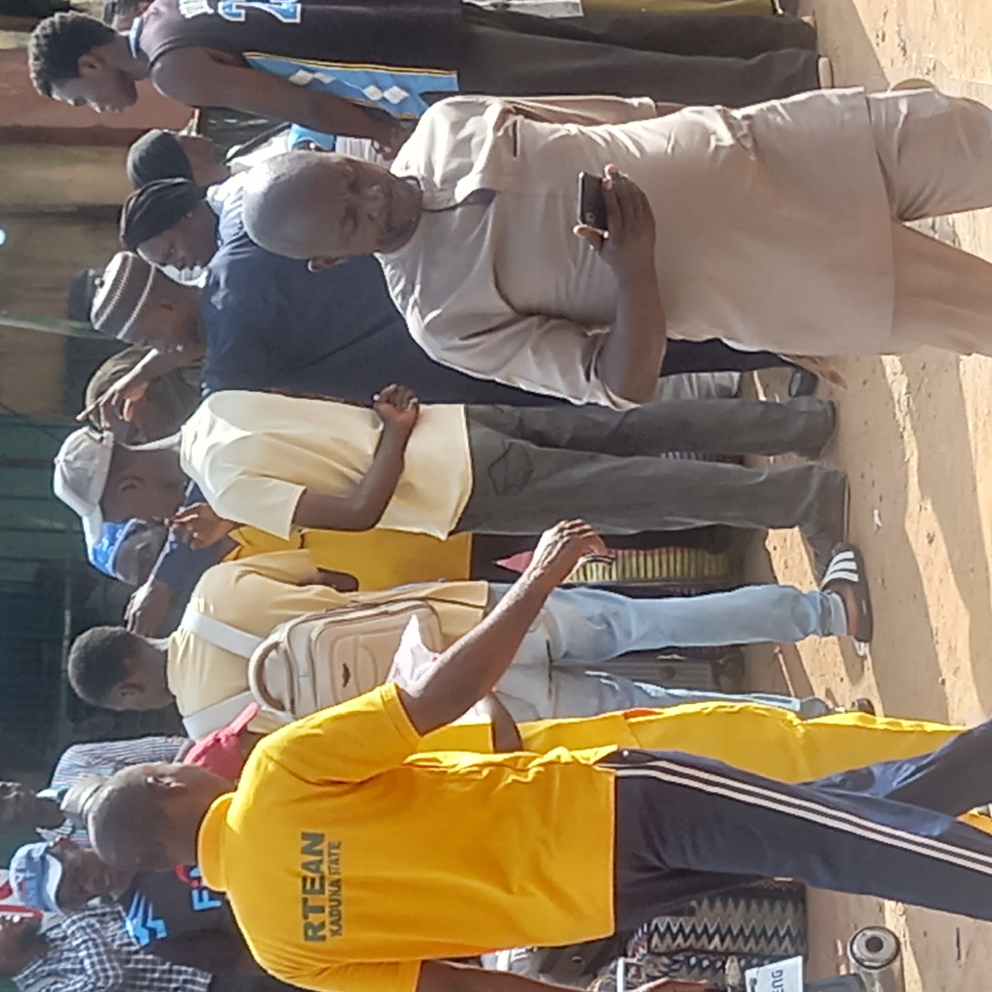 A medium shot depicts a group of approximately 12 men of varying heights and ages standing outside in Dadi Riba, Nigeria. The men are clustered closely together, with those in the foreground partially obscuring those behind. The man closest to the viewer is wearing a yellow polo shirt with the words "RTEAN KADUNA STATE" printed on the back, paired with dark blue track pants featuring white stripes down the sides. He is standing in front of a tire and an object with the text "ENG". The man to his left is wearing a light yellow shirt and has a tan satchel bag across his chest. Another man is wearing a grey shirt and pants. The background shows parts of buildings and structures, creating an urban setting. The ground appears to be dirt or a sandy color. One of the men in the group is holding a phone and appears to be taking a picture.