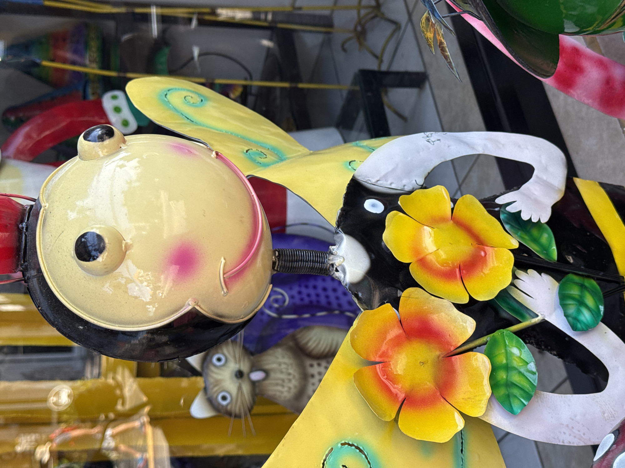 A close-up shot focuses on a whimsical, smiling bumblebee sculpture with large yellow wings, red and yellow flowers, and green leaves. The sculpture is made of painted metal, with a glossy finish reflecting the surrounding environment. In the blurred background, a quirky cat figurine with wide blue eyes and a pink tongue peeks out from behind a purple patterned surface. Further in the background, reflections in a glass window show a person wearing a hat, yellow poles, and various colorful decorative objects, suggesting a shop or market setting. The overall scene is brightly lit, with a shallow depth of field that blurs the background, emphasizing the intricate details of the bumblebee sculpture. The location is the city of Ubud District, Indonesia.