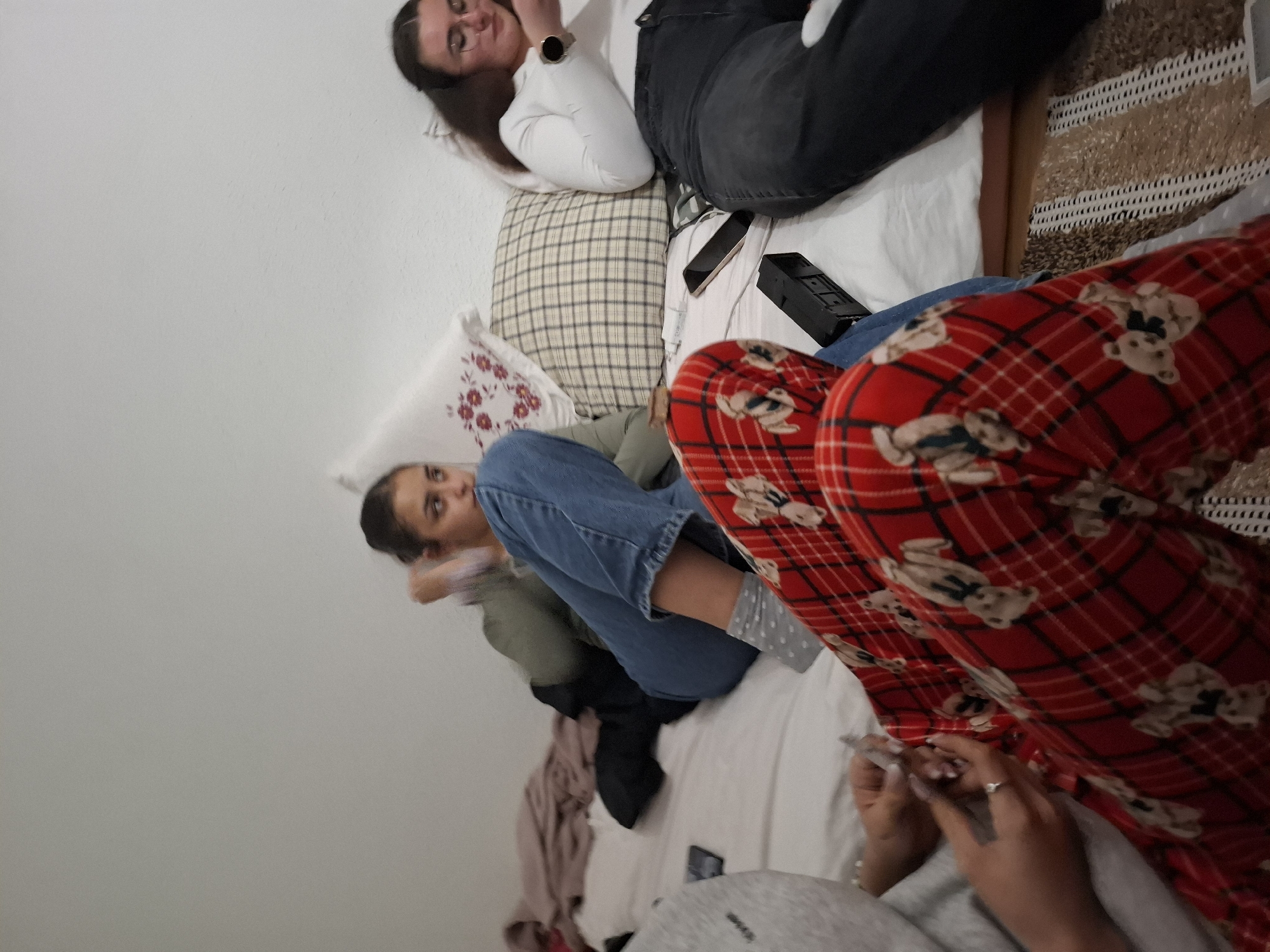 Three people are on a bed indoors. One person is wearing a white long-sleeved shirt and black jeans. The second person is wearing a green top and blue jeans. The third person is wearing red plaid pants with teddy bear patterns. Pillows with various patterns and a phone are on the bed. The location appears to be an indoor bedroom. There is no evidence to suggest that the location is in Panagyurishte, Bulgaria.