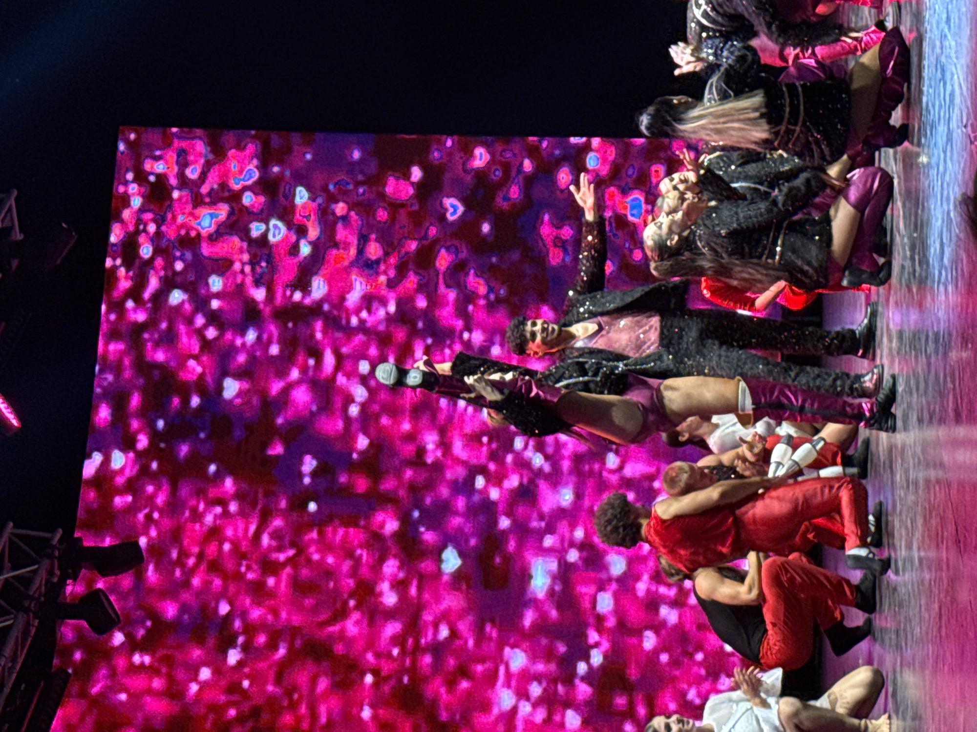 A group of dancers performs on a stage in front of a large, brightly lit pink screen displaying a pattern of hearts and bokeh effects. The screen dominates the background of the image. In the foreground, several dancers are positioned. The central figure, a dancer in a black sequined jacket and pink bodysuit, is performing a high kick with their left leg extended upwards. To their right, another male dancer in a black sequined suit and pink V-neck shirt is smiling and gesturing with his right hand. Several other dancers in red and black costumes are scattered across the stage, some crouching and some standing. The stage floor is reflective, showing the pink lighting from the screen. The overall scene suggests a performance, likely a dance show or concert, in a theatrical setting. The location is identified as Bodrum, Turkey, indicating it's a performance taking place in that city.