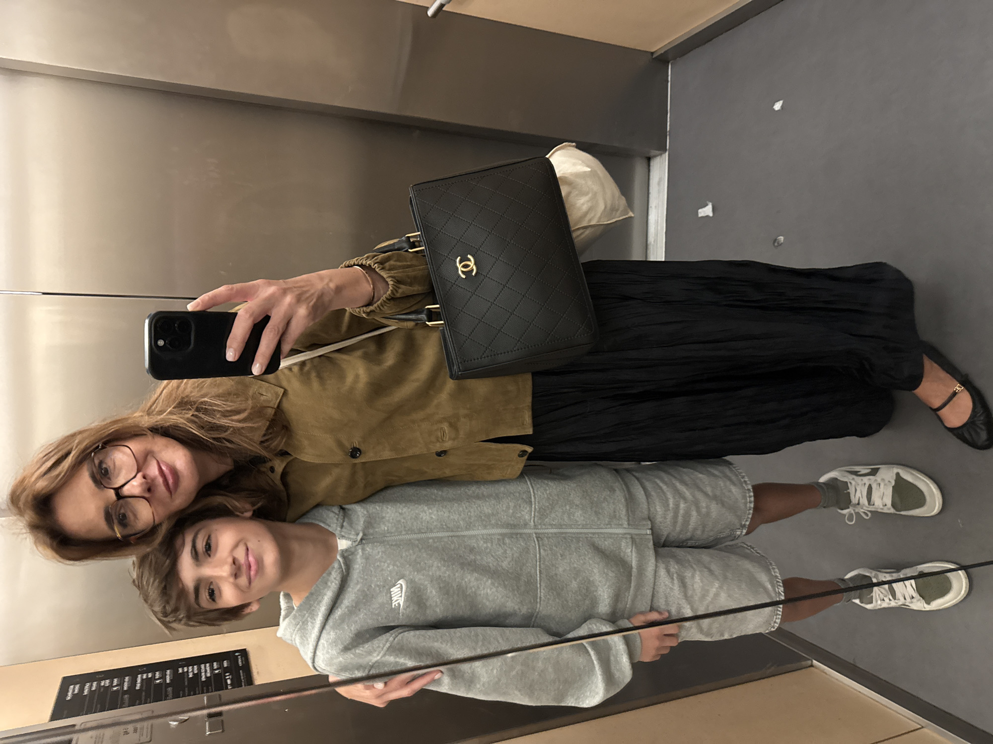 A woman and a child are in an elevator in Porto, Portugal. The woman, wearing glasses and a tan jacket, holds a smartphone to take a selfie. She carries a black Chanel purse. The child, wearing a gray Nike sweatsuit and sneakers, stands in front of her. The elevator interior is stainless steel with some signage visible. The floor is gray.