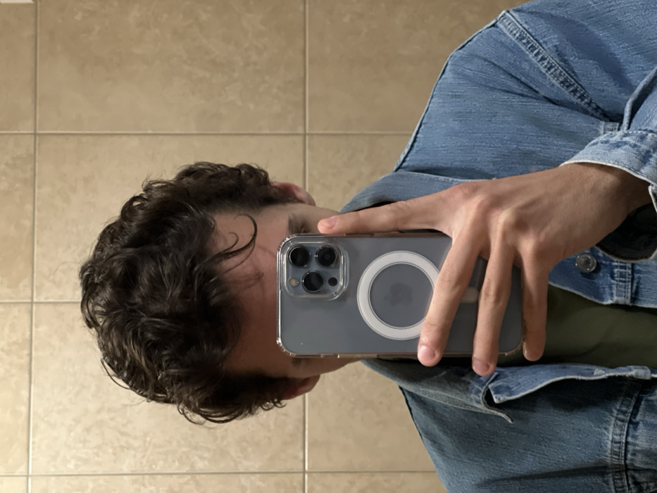 A man is taking a selfie in a bathroom mirror. His face is obscured by an iPhone with a transparent case and a magnetic ring on the back. He's wearing a denim jacket over a green shirt. The background consists of beige tiles on the wall. His hair is curly and dark. The phone is held horizontally, close to his face.