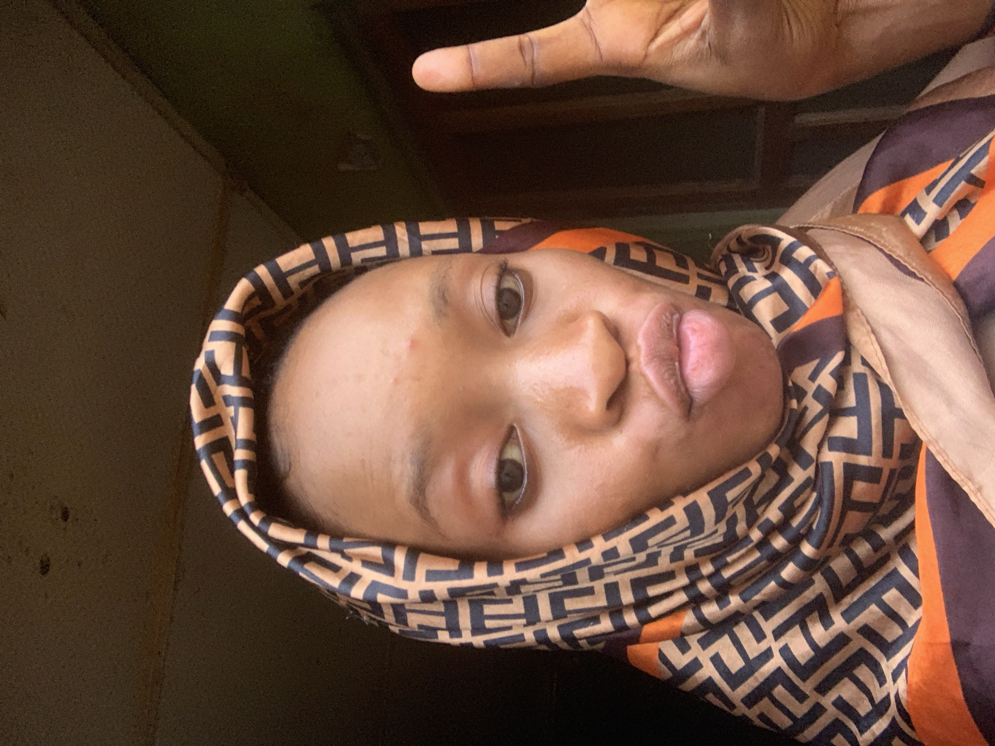 A selfie is captured from a medium close-up angle, with the subject, a young woman, in the foreground. She is wearing a patterned headscarf with a repeating geometric design in black and brown on an orange background. Her face is turned slightly towards the camera, with her lips pursed and her right hand raised in a peace sign. The background is dimly lit and appears to be indoors, with a dark, textured ceiling and a hint of green on the right side, suggesting a wall or an opening to the outside. The overall impression is of a candid moment captured in a simple, unadorned setting, possibly in Shagamu, Nigeria, given the context.