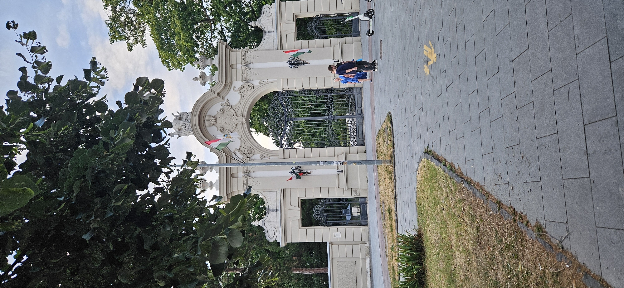 The image shows a large, ornate archway, which is identified as the entrance to the Festetics Palace in Keszthely, Hungary. The archway is made of light-colored stone and features intricate carvings and decorative elements, including statues and urns. Two Hungarian flags are prominently displayed on poles flanking the central arch, one higher and one lower on the right. Behind the flags, the flags of Hungary are visible.
In front of the archway, there are two sets of large, decorative iron gates, which appear to be closed. To the left of the archway, a smaller, similarly styled iron gate is visible within a recessed section of the wall.
On the paved walkway in front of the archway, three people are visible. One person, wearing a blue dress, is standing near the right-side gate. Another person, wearing a dark shirt and pants, is standing a short distance away, holding a phone. A third person, a child, is partially visible behind the person in the blue dress.
To the right of the archway and people, a white electric scooter is parked on the sidewalk. The ground in front of the archway is a paved walkway with gray rectangular stones. To the left of the walkway, there is a strip of dry, yellowing grass with some green plants.
The scene is framed by the dark green leaves and branches of a large tree in the foreground, which partially obscures the top of the archway and the sky. The sky is partly cloudy with a soft, diffused light, suggesting it is either early morning or late afternoon. The overall impression is of a grand historical entrance on a pleasant day.