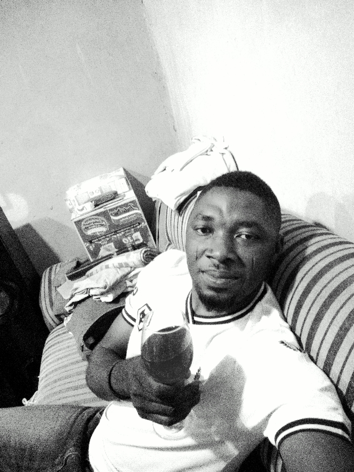 A black and white image displays a male individual, appearing to be an adult of African descent, seated indoors in Gwagwarwa, Nigeria. The man, positioned with a slight smile and direct gaze towards the camera, has short dark hair and a beard. He wears a light-colored polo shirt with dark stripes on the collar and sleeve cuffs, and dark trousers or jeans. His left hand holds a stemmed glass containing a dark liquid. He is leaning against a striped sofa or cushion. In the background, to his left, is a corner formed by two plain, light-colored walls. A carton, likely a product box with visible branding resembling "Minimie," is situated on a surface behind him, alongside various folded fabric items and a rolled textile object.