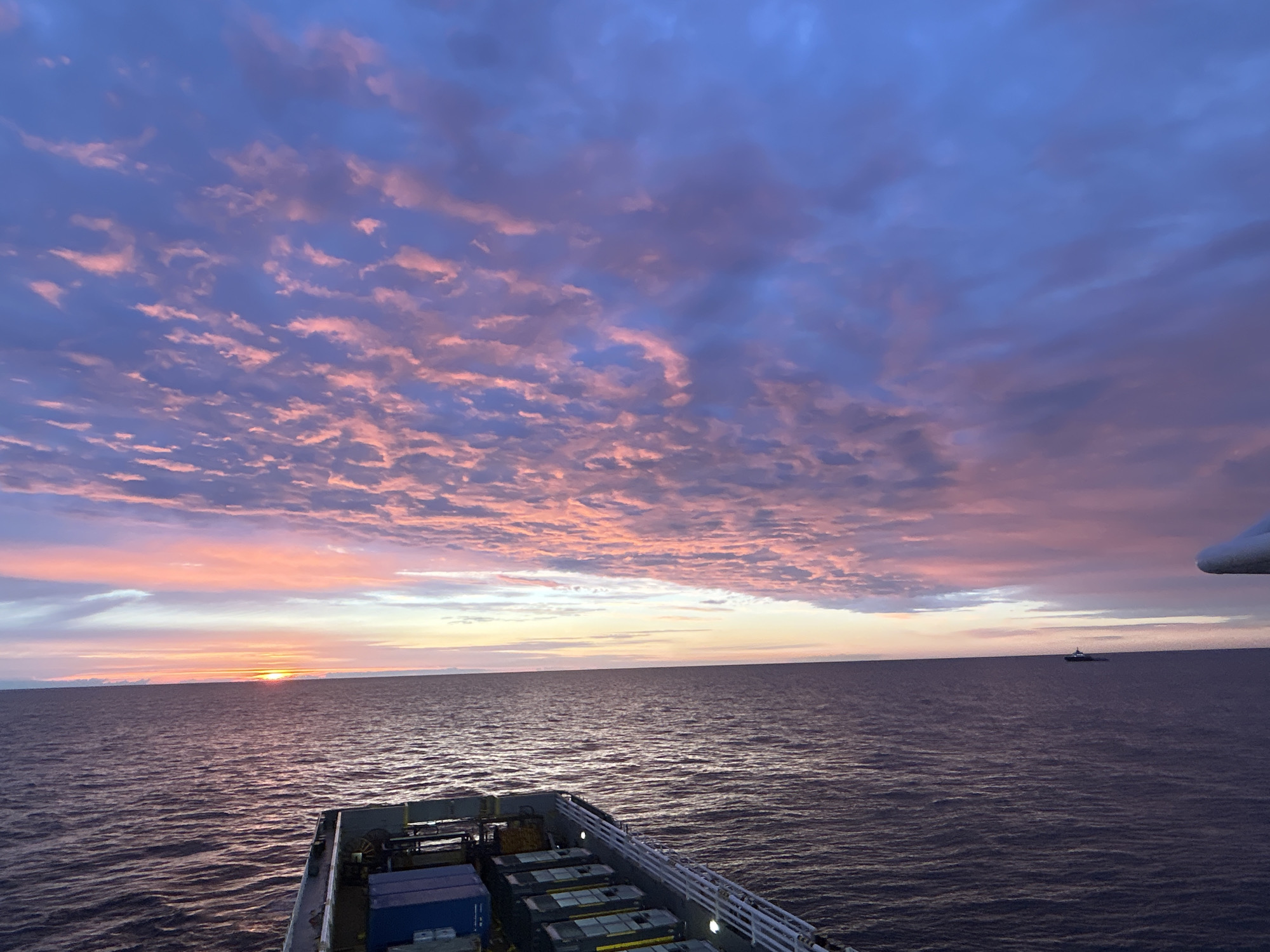 An outdoor, wide shot captures a vast ocean under a dramatic sky filled with colorful clouds during what appears to be sunrise or sunset. The sun, a bright orange disc, is low on the horizon, casting a warm glow across the water. The sky is a gradient of blues and purples, with streaks of pink and orange illuminating the clouds. In the foreground, the deck of a ship is visible, loaded with stacked shipping containers, some blue and others dark. The ship is a dark silhouette against the vibrant sky. In the distance, a small boat can be seen on the water, a solitary speck on the expansive seascape. The overall scene evokes a sense of tranquility and the immense beauty of nature.