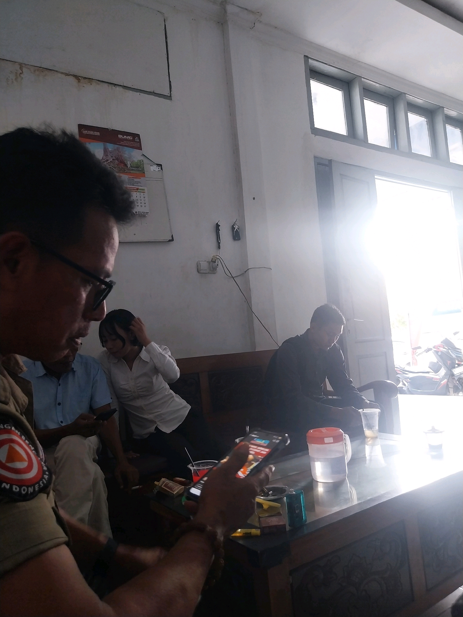 The image depicts an indoor scene likely within a public building in Nganjuk, Indonesia. In the foreground, a person wearing a tan uniform with a circular patch featuring a red triangle and white border is visible from the chest up. They are holding a smartphone and appear to be looking at it. To their left, a man in a blue shirt is seated and also looking at a smartphone. Behind them, a woman in a white blouse is touching her hair and looking down. Further back, a man in a black long-sleeved shirt is seated, looking down at a table. The environment is furnished with wooden seating and a table. On the table are various objects including a pitcher of water, a plastic cup, cigarette packs, and a vape device. Sunlight streams in from an open doorway or large windows on the right side of the image, creating a bright glare. The walls are white, and a calendar with a "BUMD" logo is visible on the left wall. The overall impression is a casual gathering or waiting area.