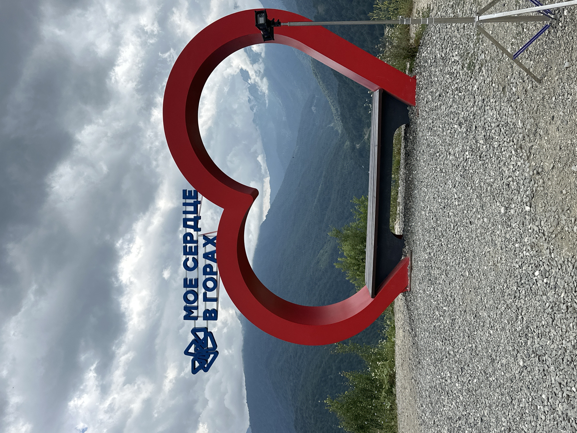 An outdoor, eye-level, medium shot shows a large red heart-shaped bench with the inscription "МОЕ СЕРДЦЕ В ГОРАХ" (My Heart in the Mountains) in blue Cyrillic lettering in the city of Estosadok, Russia. The bench is situated on a gravelly surface overlooking a vast mountainous landscape. The sky above is overcast with dramatic grey clouds. A tripod with a camera is positioned to the right of the heart-shaped bench, suggesting it is set up for photography or videography. Green foliage and white wildflowers grow around the base of the bench and the surrounding area. The mountains in the background are densely forested and shrouded in mist, creating a sense of depth and scale.