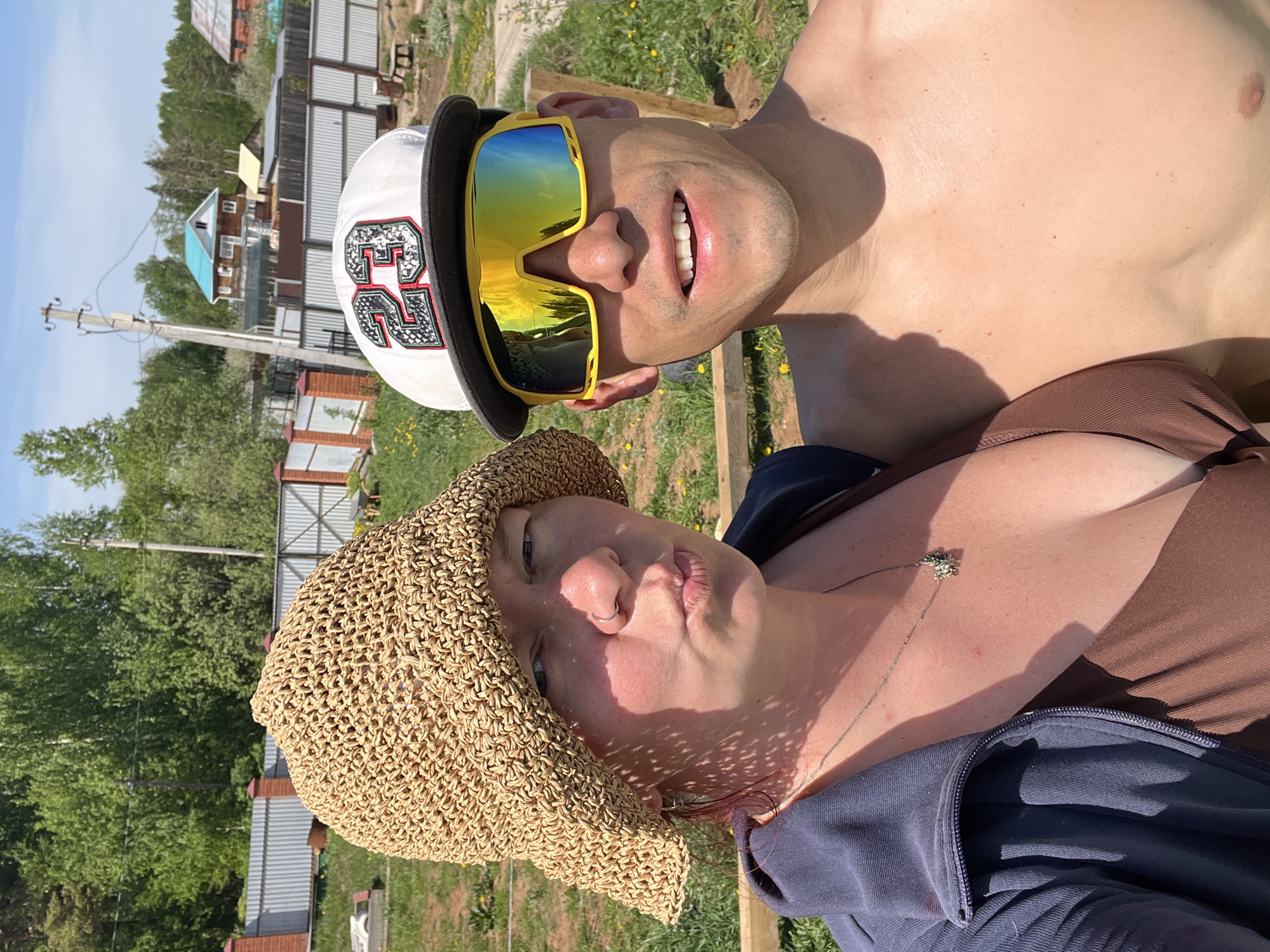 A selfie of two people outdoors during daylight. The male is shirtless, wearing yellow sunglasses and a white cap with the number "23" on it. He has a wide smile. The female is wearing a woven sun hat, a brown top, and a blue jacket. She has a necklace with a pendant and is making a kissing face. The background shows houses, trees, and grass, suggesting a rural or suburban setting. The exact location in Chaikovsky, Russia, cannot be determined from the image alone.