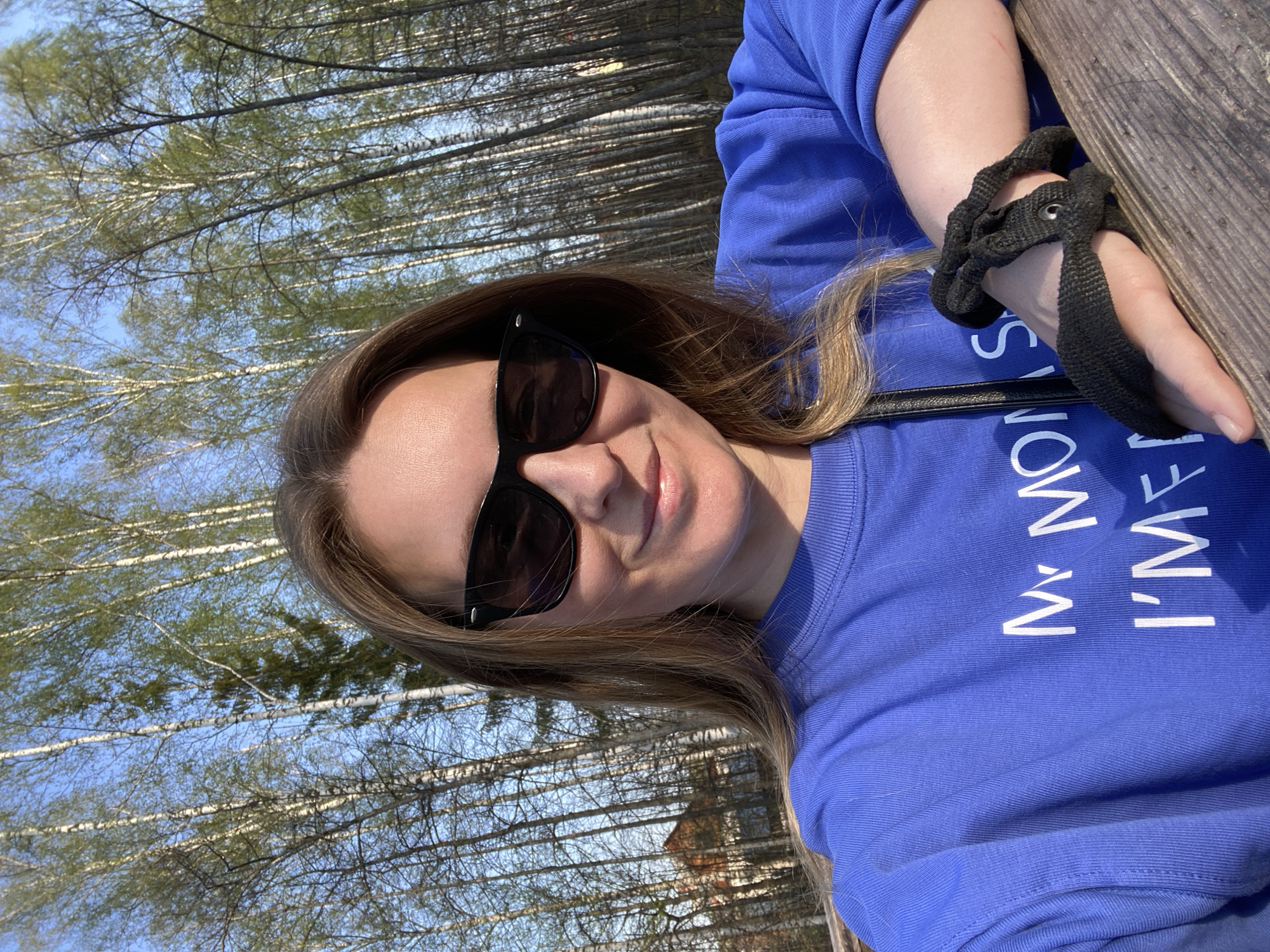 A woman with long brown hair and sunglasses is the central subject of the image. She is wearing a blue sweatshirt with white text. A black strap-like object is wrapped around her hand and a wooden structure. In the background, numerous tall, thin trees with white bark are visible. Patches of blue sky are interspersed among the trees. Part of a building with a brown roof is seen between the trees in the mid-ground. Based on the landscape and architecture, the location is likely in Noviy, Russia.