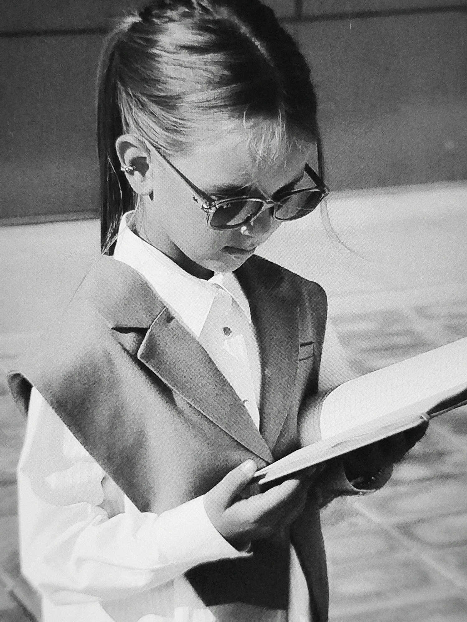 A black and white, medium shot captures a young girl wearing sunglasses and a suit, reading a book. She is positioned in what appears to be an outdoor, possibly urban setting, given the textured ground and indeterminate background suggesting a cityscape. Her hair is pulled back in a ponytail, and she has an earring in her left ear. The girl is dressed in a white collared shirt, a vest, and a blazer, giving her a professional appearance. Her gaze is directed downwards at the open book she holds in her hands. The lighting casts subtle shadows, highlighting the details of her clothing and the book.