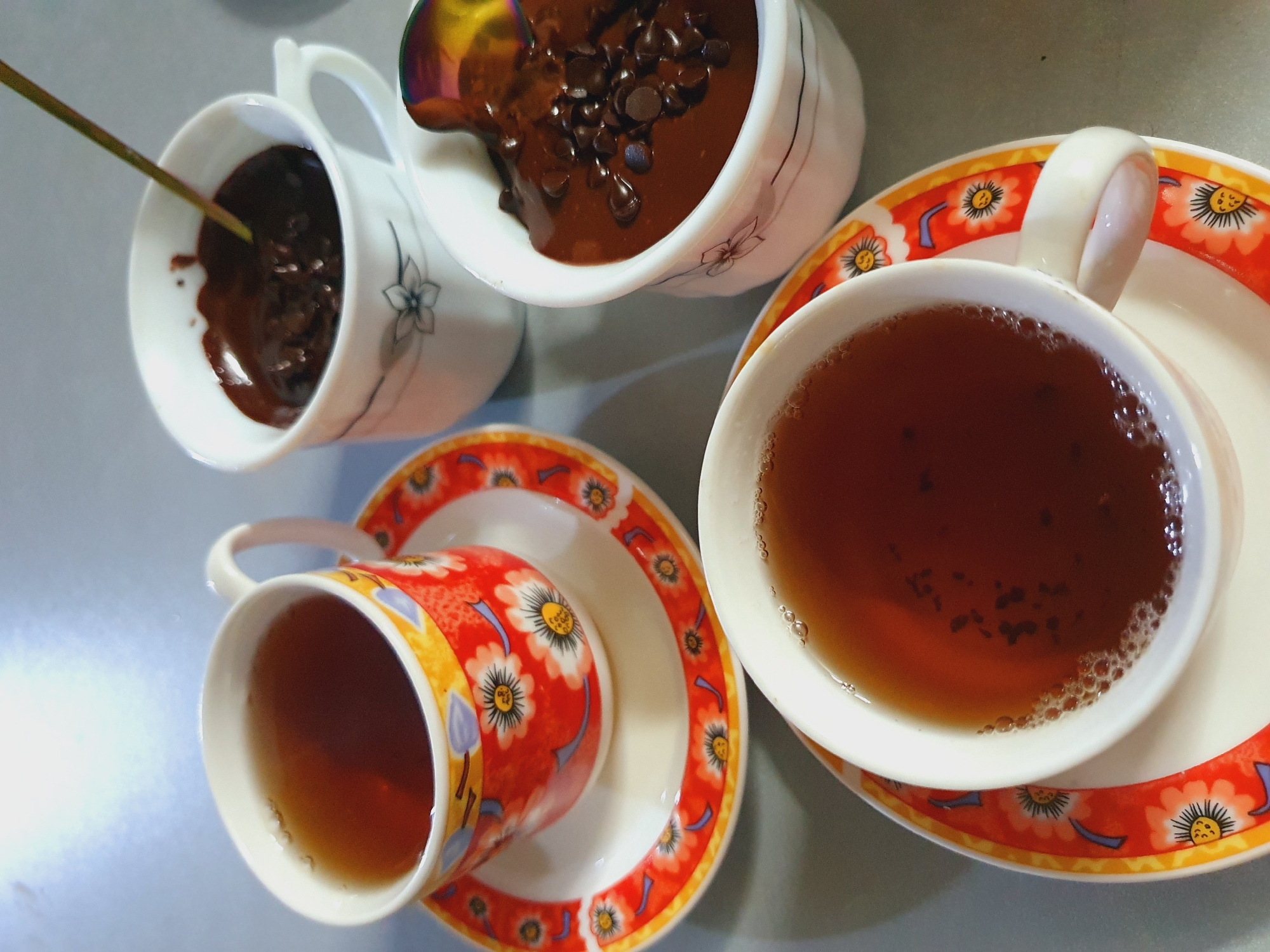The image depicts a close-up, overhead view of a table setting with three cups, two filled with hot chocolate and one with tea. The cups are arranged on a gray surface, possibly a tabletop. One cup of hot chocolate is in the foreground, a white ceramic cup with a subtle floral design, filled with a dark brown liquid and topped with chocolate chips. A spoon with a multicolored, iridescent finish is partially submerged in the hot chocolate. To the left of this cup, another white ceramic cup, also with a floral design, contains hot chocolate and has a thin, light-colored stick inserted into it. In the upper left quadrant, a tea cup with a vibrant red and orange floral pattern sits on its saucer, filled with amber-colored tea. A second tea cup, similar in design to the first, is partially visible in the lower portion of the image, also on its saucer and filled with tea. The lighting suggests an indoor setting, possibly a home environment in Shiraz, Iran, given the floral patterns on the cups which are reminiscent of traditional Iranian ceramics. There are no people visible in the image, and the focus is solely on the beverages and their presentation. The scene appears to be set for a relaxed gathering or a moment of personal indulgence.