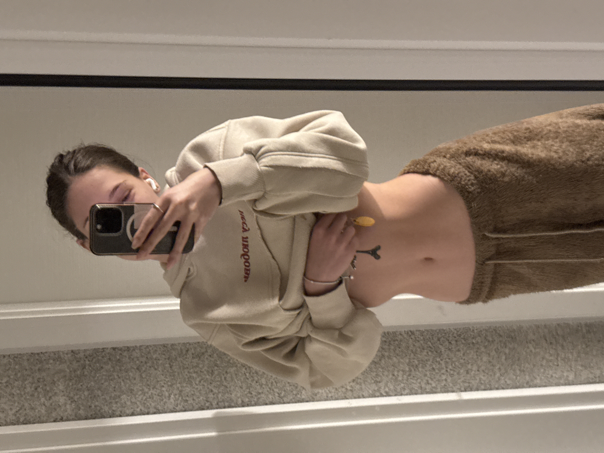 A slender person with dark hair takes a selfie. The person wears a loose, light tan hoodie that is partially lifted to reveal their midriff, which features a small Eiffel Tower tattoo. They are also wearing brown, fuzzy sweatpants. The person is facing left, holding a phone in front of their face. The background shows a white wall with a textured carpeted floor. There is no indication of the location in Sofia, Bulgaria.