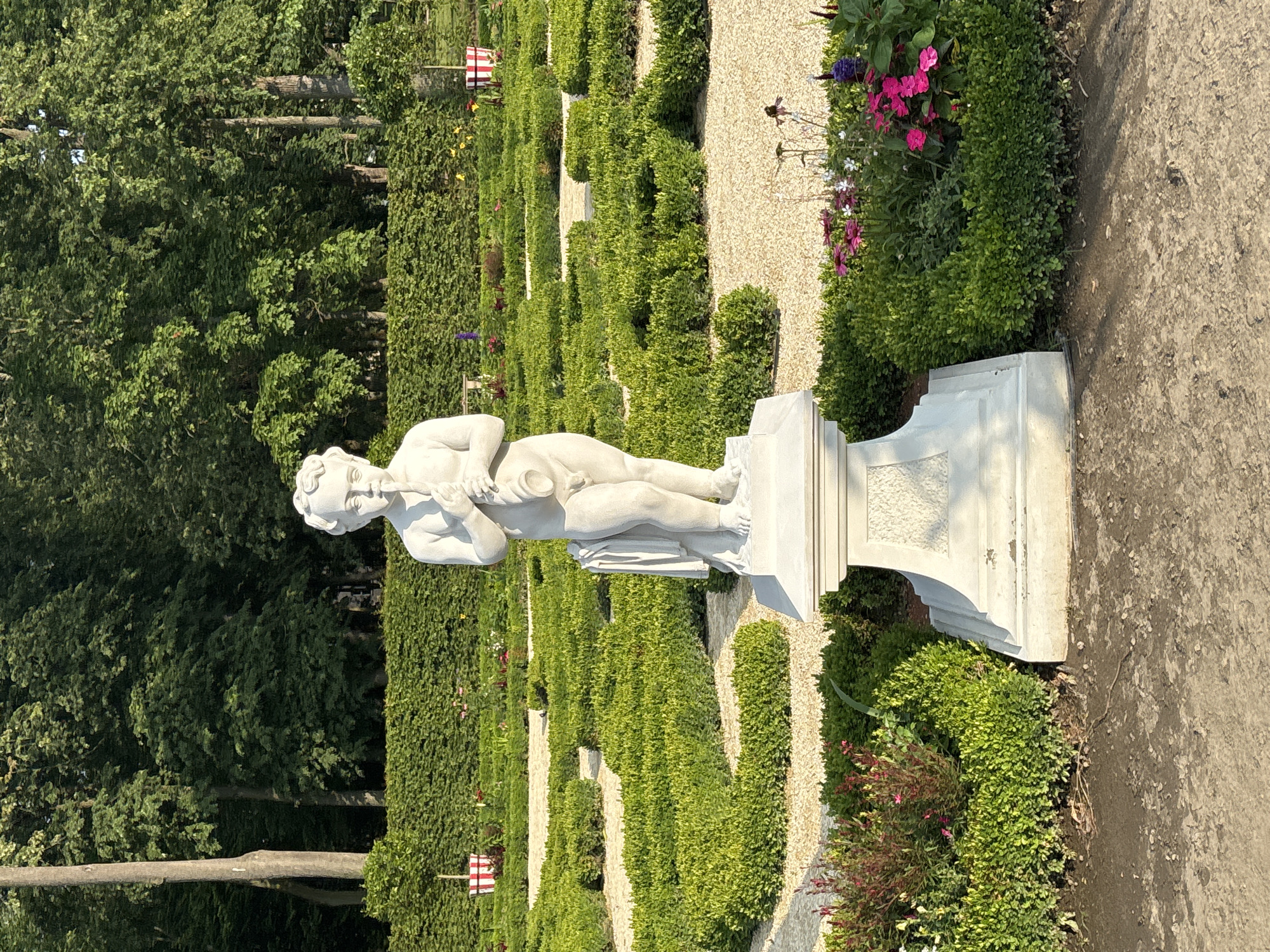 The image shows a white stone statue of a young boy holding a flute, positioned on a pedestal in the center of a formal garden. The garden features meticulously manicured green hedges arranged in a geometric pattern, interspersed with gravel pathways. In the background, a dense line of tall green trees forms a natural backdrop, with a clear blue sky visible above. The scene is well-lit, suggesting it was captured during daylight hours, likely in the morning or late afternoon due to the soft quality of the light and the shadows.
This image depicts the Sunken Garden located in Branicki Palace in Białystok, Poland. The Sunken Garden is a significant part of the palace's baroque complex, known for its preserved historical layout and ornamental greenery. The statue of the boy with the flute is a typical element found in such formal gardens, often symbolizing music or youth. The overall atmosphere is serene and well-maintained, reflecting the historical and aesthetic significance of the location.