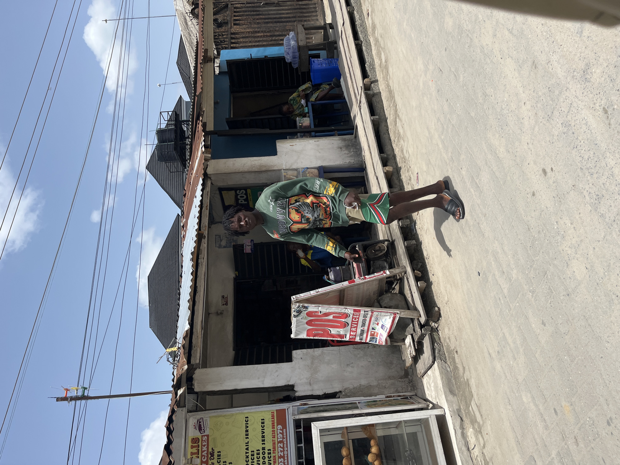 A person stands on a sidewalk in front of a building in Iranla, Nigeria. The person is wearing a green shirt with a print design and green and red shorts, and sandals. A sign that reads "POS SERVICES" is placed near the individual in front of a building that also has another sign reading "ELIS CAKES", indicating local businesses. The sidewalk is cracked and aged. Overhead, electrical wires are visible against a partially cloudy sky. In the background, another individual sits indoors on a chair.