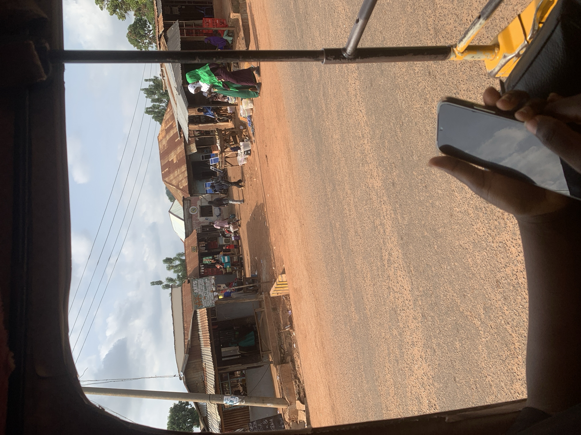 The image captures a street scene in Lafia, Nigeria, as viewed from inside a vehicle, likely an auto-rickshaw. The perspective is tilted, suggesting movement. The foreground is dominated by the interior of the vehicle, with a brown frame on the left and a portion of a yellow and black structure on the right, possibly part of the vehicle's framework. A hand holding a smartphone is visible on the right side, with the screen reflecting a cloudy sky.
The scene outside the vehicle features a dusty, unpaved road running parallel to a row of shops and market stalls. The buildings are made of corrugated metal and wood, with some showing signs of wear and tear. Several people are visible, some standing or walking near the shops, while others appear to be engaged in activities within the market. The shops display various goods, including what appear to be packaged items and textiles. A woman in a bright green headscarf and dark dress is a prominent figure among the shoppers. Power lines crisscross the sky above the street.
The weather appears to be partly cloudy, with patches of blue sky visible between the clouds. The light suggests it is daytime, possibly late morning or early afternoon. The overall atmosphere seems to be that of a bustling local market in a developing area. A sign is partially visible on one of the stalls with text that includes "ARTS" and possibly other words in a local language. Another sign has what looks like a date, "#10-25". The unpaved road and the style of the buildings are strong indicators of the location in Nigeria.