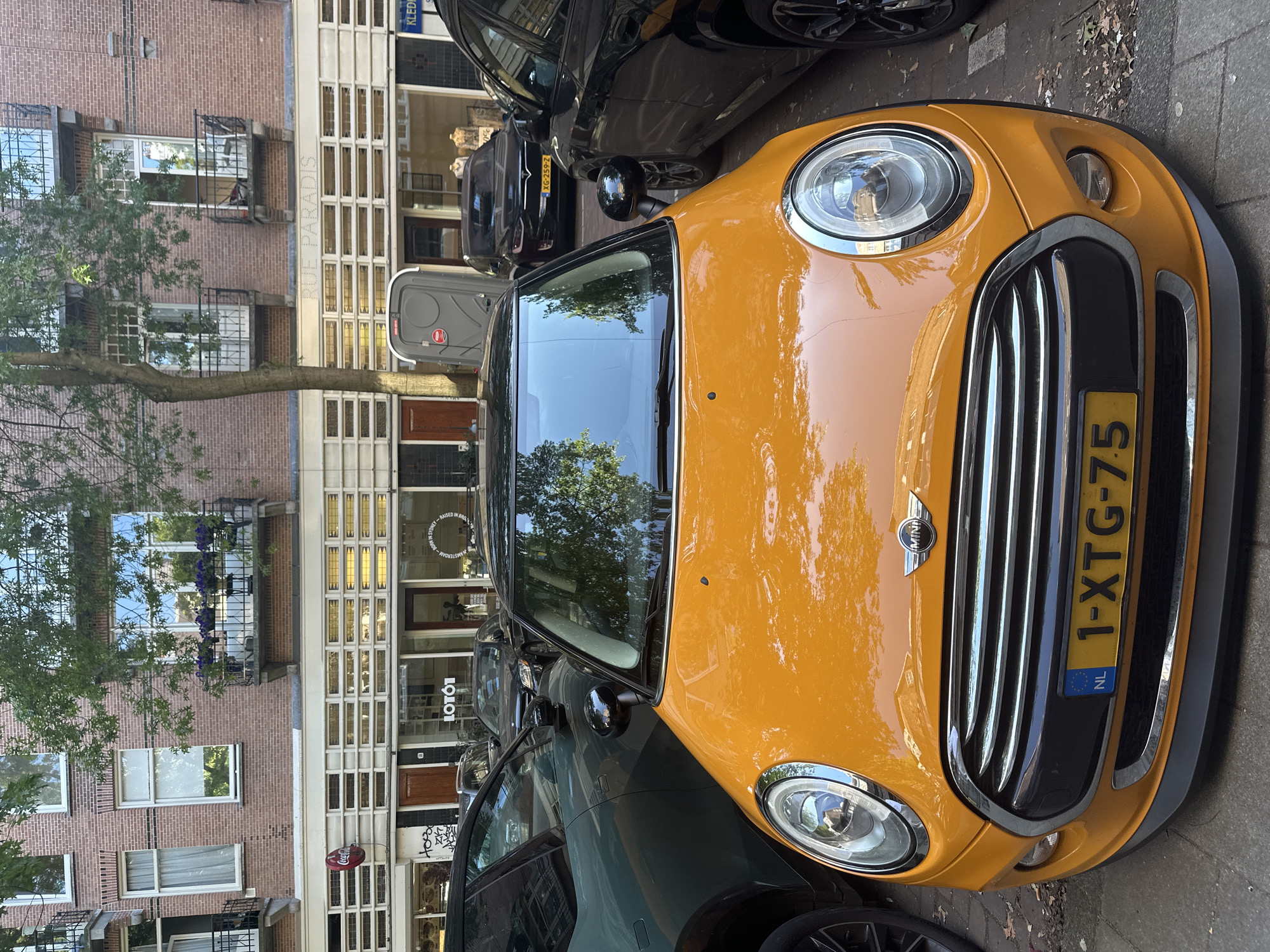 In Amsterdam, Netherlands, an orange Mini Cooper is parked on a street in front of a brick building. The Mini Cooper has a black roof and a Dutch license plate that reads "1-XTG-75". To the left of the Mini Cooper is a dark green SUV, and to the right is a black SUV. The brick building behind the cars has several windows, some with balconies, and a sign that says "RUE PARADIS". There is also a portable toilet on the sidewalk near the building entrance. The scene is captured in daylight, with trees providing dappled shade on the cars and sidewalk.