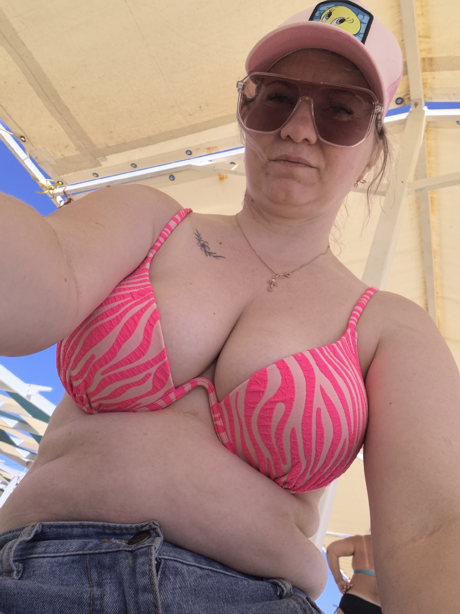 A woman is pictured in a selfie taken outdoors. She is wearing a pink and beige striped bikini top, denim shorts, sunglasses, and a pink baseball cap with a cartoon character on it. She has a tattoo of a flower on her left shoulder and a necklace with a cross pendant. The background shows part of a beach umbrella and a blue sky. The image appears to have been taken in Anapa, Russia, a city known for its beaches and resorts.