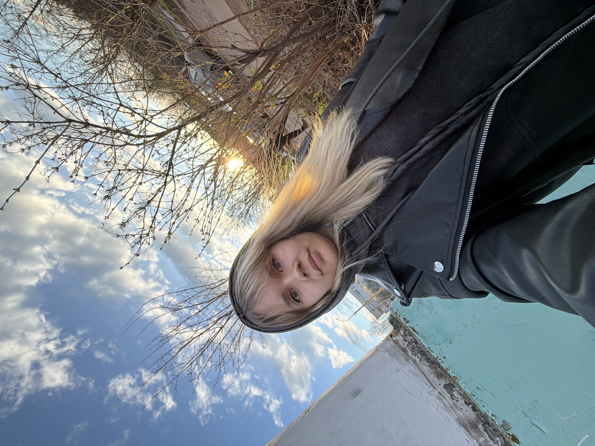 A rotated selfie captures a young woman outdoors during what appears to be golden hour. The woman, with blonde hair and fair skin, is looking directly at the camera with a neutral expression. She wears a dark, possibly black, hooded jacket with a visible zipper and a silver snap button. Her blonde hair is highlighted by the warm sunlight.
The background features a clear blue sky with scattered white clouds, suggesting good weather. Bare tree branches with small, emerging buds are prominent, indicating either late winter or early spring. The sun is visible in the upper right quadrant of the image, casting a warm, bright glow. Beyond the trees, a road or path and part of a car are faintly visible. In the foreground, below the woman, there's a light-colored wall or structure with a distinct turquoise-painted top edge, showing some signs of wear and peeling. No visible text is present in the image. The overall impression is a sunny outdoor moment in a natural or semi-urban setting.