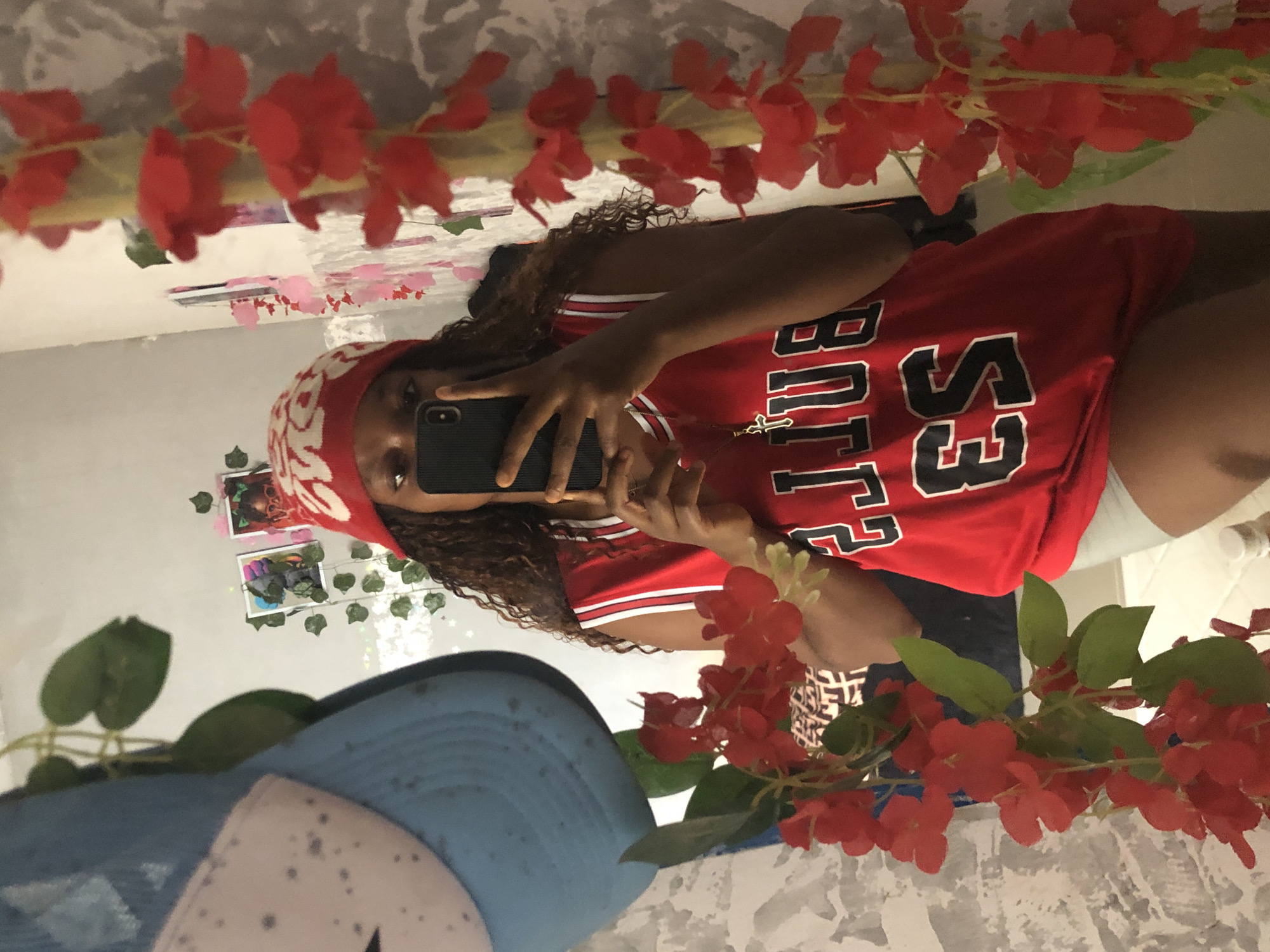 The image shows a person taking a selfie in a mirror. The person is wearing a red jersey, a red and white head covering, and a gold necklace with a cross. They are holding a dark-colored smartphone. The background includes red flowers, a blue and white baseball cap, and photographs on the wall. The scene appears to be indoors, likely in a bathroom or bedroom based on the presence of a mirror and wall decorations. The location is unknown, and there is no evidence in the photo to suggest it is in the city of Kashere, Nigeria.