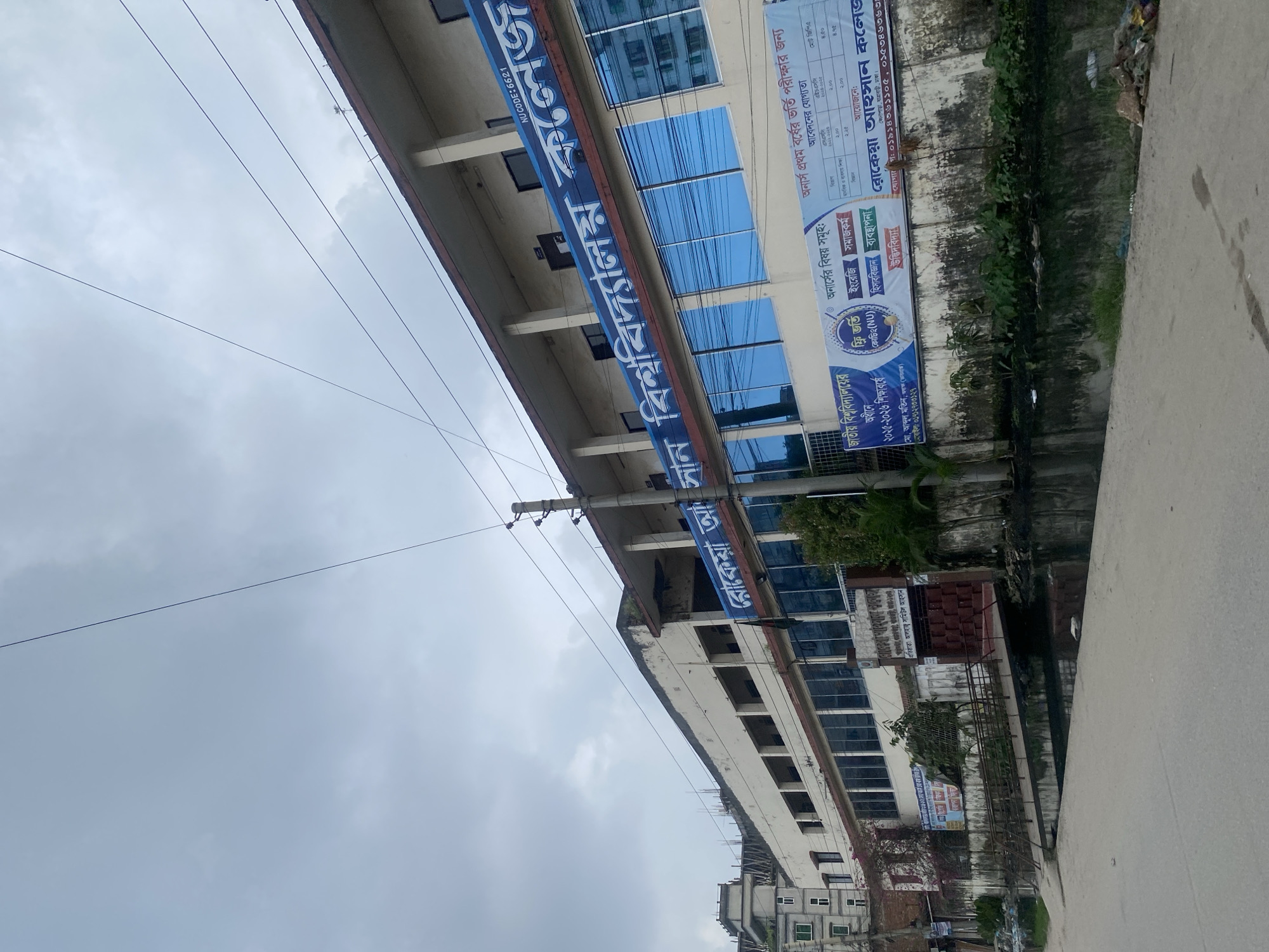 The image shows a large, multi-story building with blue-tinted windows. A banner across the front of the building displays text in Bengali, indicating it is a college. The building appears to be in an urban environment, with a street and a canal in the foreground. There are some electrical wires running across the sky. The sky is overcast with clouds. The overall impression is that of a college campus or a large educational institution in a city. The banner indicates the college is affiliated with the National University and mentions admissions for the academic year 2021-2022. It also lists various subjects like English, Economics, Social Work, History, Sociology, and Business Administration. The building's architecture suggests it's a modern structure, possibly built in the late 20th or early 21st century. The presence of the canal and some visible garbage suggests the area may not be as well-maintained as a typical pristine campus. The building's facade has some signs of wear and tear, with peeling paint visible on the upper sections. The image is taken from a slightly low angle, emphasizing the height of the building.