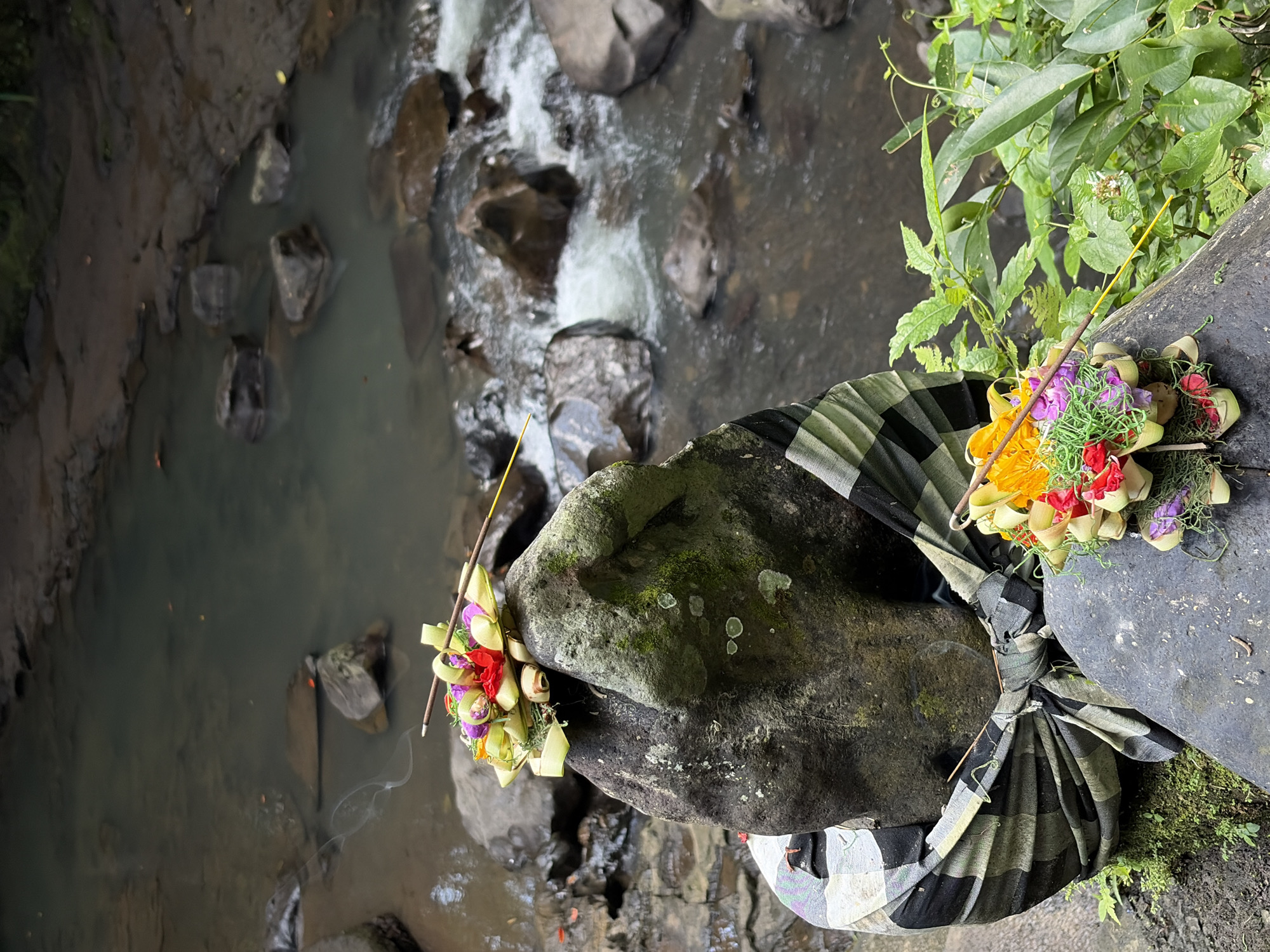 This image depicts a spiritual offering at a riverbank in Gianyar, Indonesia. A large, dark rock, wrapped in a traditional black and white checkered Balinese sarong (kain poleng), stands prominently in the foreground. Atop this rock rests a small, meticulously arranged floral offering in a woven leaf basket, adorned with red, pink, and purple flowers, and a single lit incense stick emitting a thin trail of smoke. Another similar offering is placed on a smaller rock in the lower right corner of the frame, featuring yellow and red flowers. The background is a flowing river with submerged rocks, its water appearing murky and dark. Lush green foliage surrounds the scene, suggesting a natural, possibly sacred, outdoor location. No people are visible in the image.